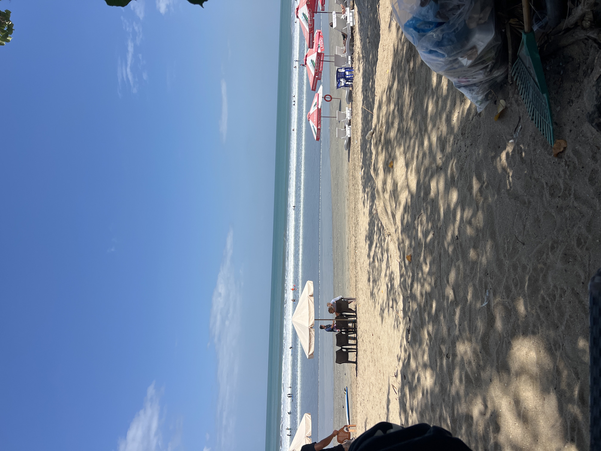 This is a wide shot of Kuta Beach, Indonesia, on a sunny day. The sand is a light brown, and there are several beach umbrellas and lounge chairs set up. In the foreground, there is a large pile of trash bags and a rake. In the middle ground, there are a few people sitting at tables under an umbrella, and several people are in the ocean, some of whom appear to be surfing. The ocean is a vibrant blue-green, with white waves breaking on the shore. The sky is a clear, bright blue with a few scattered white clouds. The overall scene depicts a typical beach day, with a hint of the ongoing effort to keep the beach clean.
