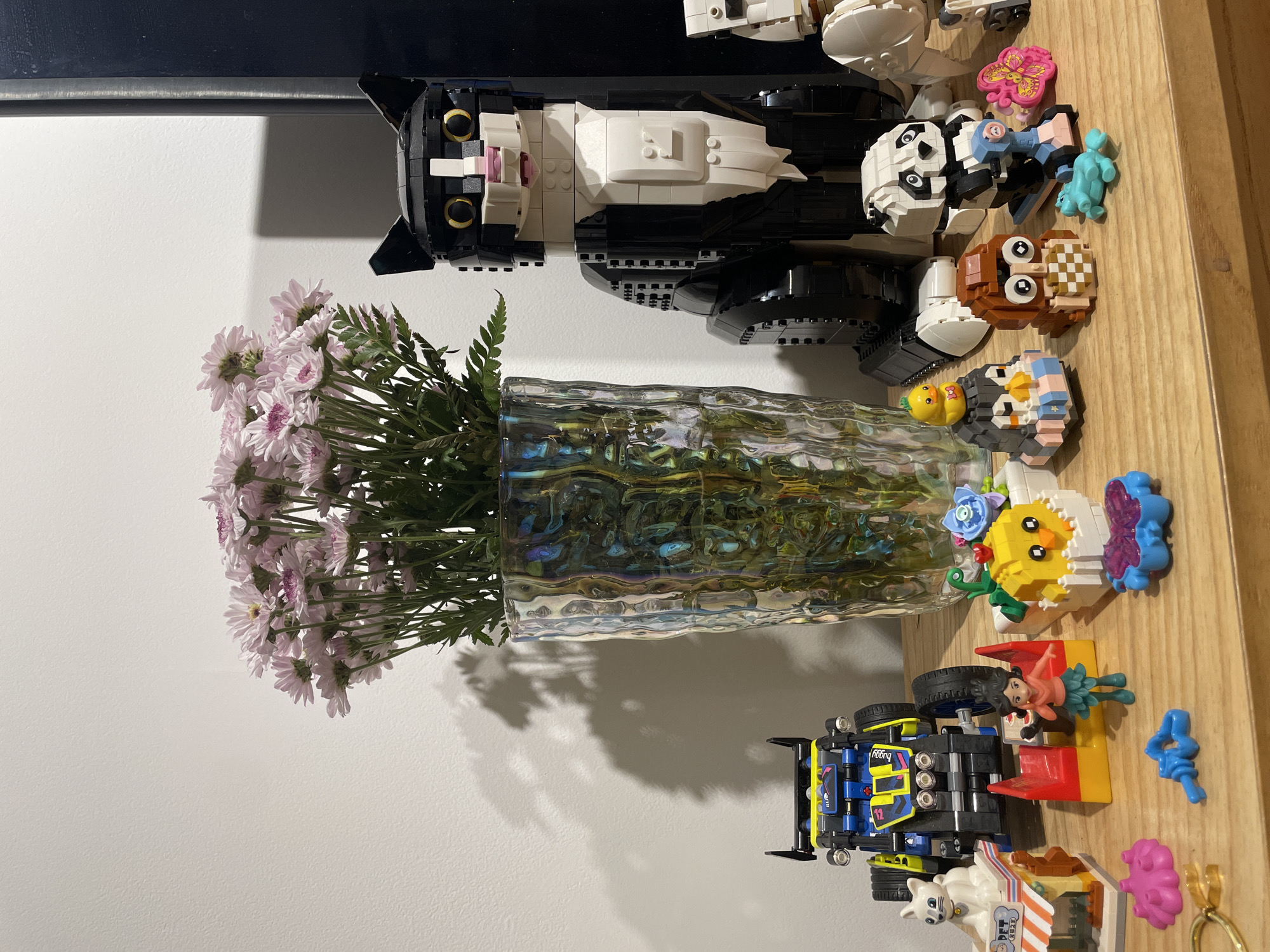 The image shows a still life arrangement on a wooden surface. From top to bottom: a Lego cat, followed by a Lego panda, a Lego owl, and other small Lego creations. Below the Lego creations is a glass vase containing flowers. Further down, is a Lego race car with a minifigure, and more Lego creations. The background consists of a white wall and a dark-colored shelf or frame above. There's no indication of the location being in Buenos Aires, Argentina, or of any human presence or interaction.