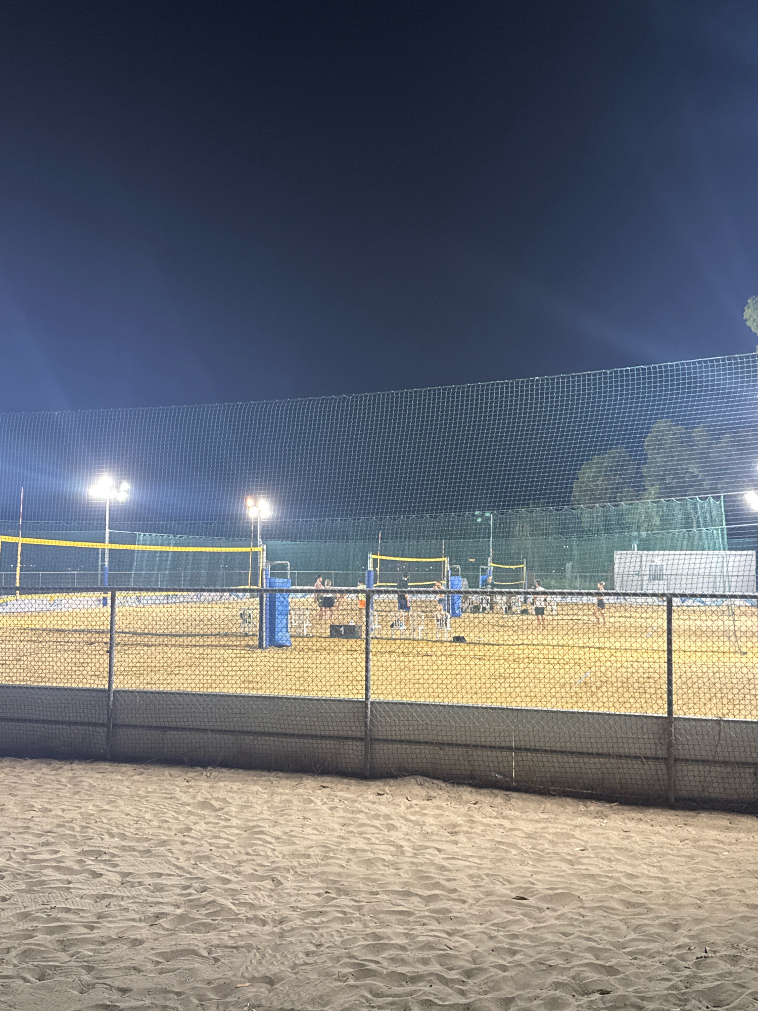 The image depicts an outdoor beach volleyball court at night, illuminated by floodlights. Several people are visible on the court, engaged in playing volleyball. The court is enclosed by a fence, with sand as the playing surface. In the foreground, a portion of another sandy area is visible, possibly a seating area or another court. The background shows trees and a dark sky. The scene appears to be in Limassol, Cyprus, a coastal city known for its beaches and recreational facilities.