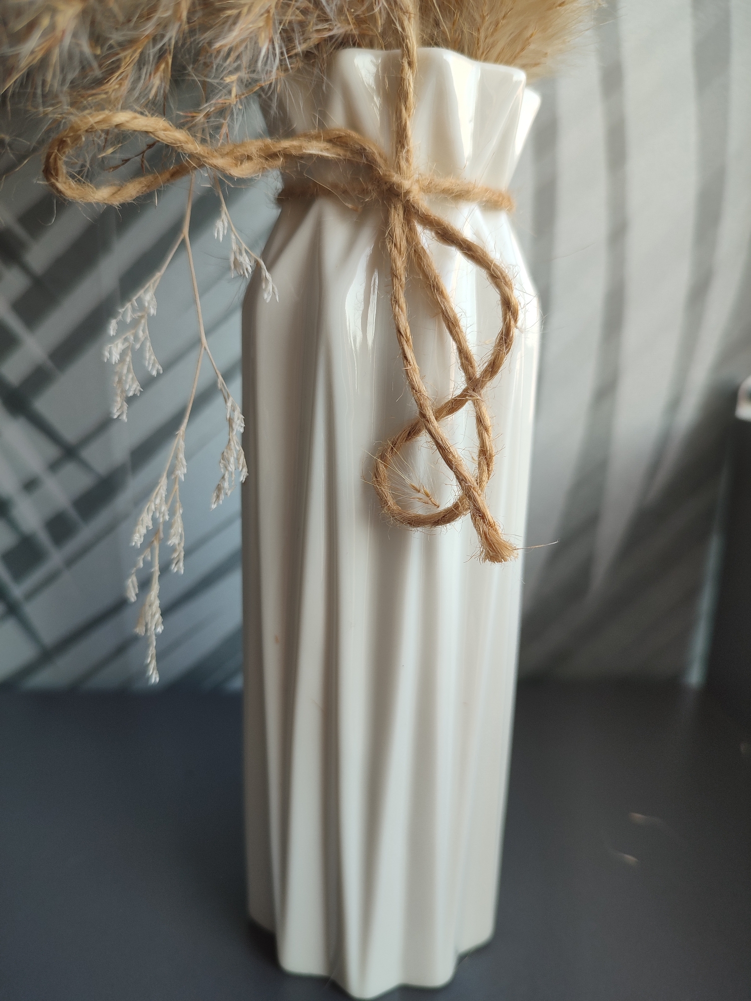 A white, ribbed ceramic vase with dried decorative grasses is placed on a dark gray surface. The vase is tied with rough, brown twine. A delicate, dried white sprig hangs from the twine. The background consists of a gray surface with a diagonal striped pattern, possibly a wall or blinds, and out-of-focus vertical lines. No people or any recognizable landmarks of Voronezh, Russia are visible.