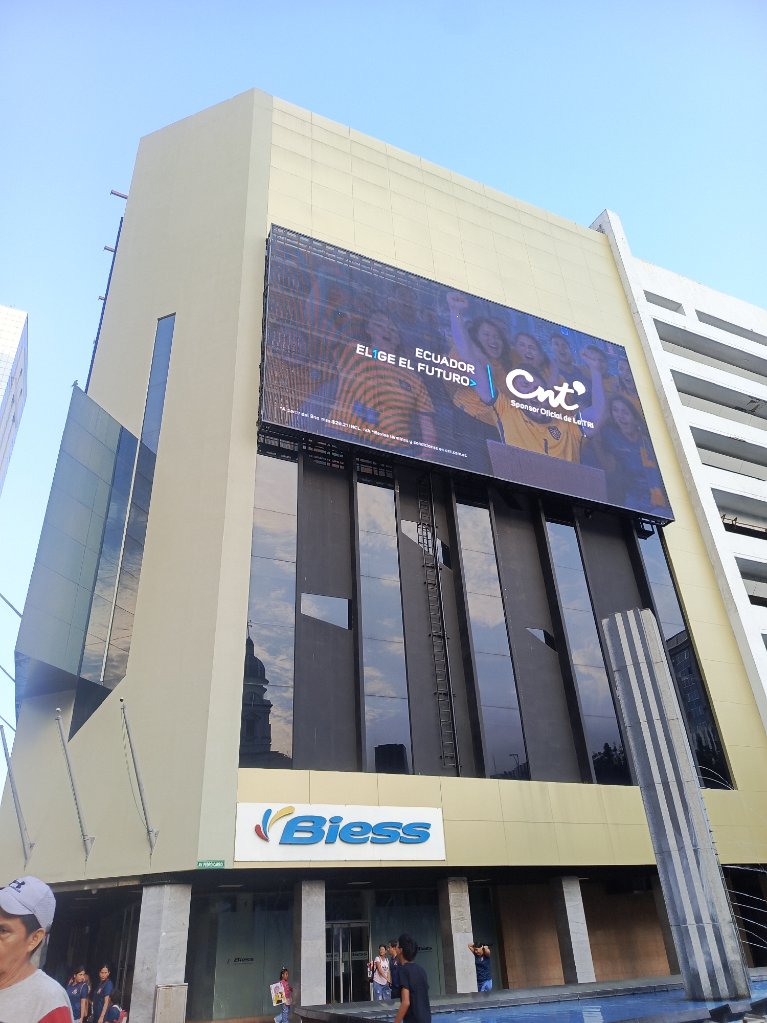 This image shows a modern building in Guayaquil, Ecuador, featuring a large digital billboard displaying an advertisement. The billboard shows the words "ECUADOR ELIGE EL FUTURO" and the logo of "Cnt Sponsor Oficial de La Tri". Below the billboard, the building has large, dark tinted windows and a sign that reads "Biess". The lower part of the building has an entrance area with a reflective pool in front. Several people are visible on the sidewalk in front of the building, some are walking and others appear to be congregating. The sky is clear blue.