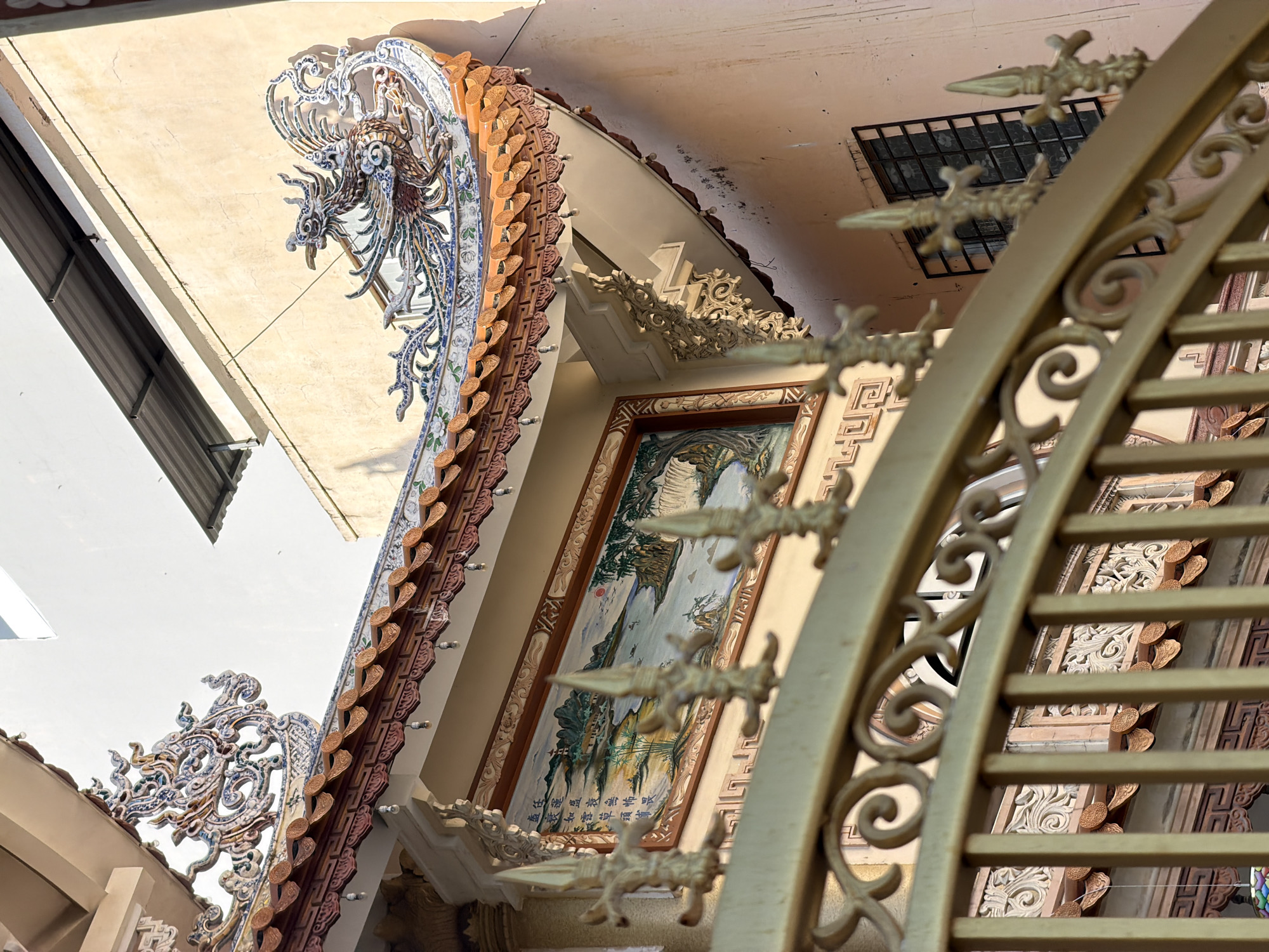 This image depicts architectural details of a building in Tay Nha Trang Ward, Vietnam. The structures display ornate, traditional Vietnamese ornamentation. Key elements include tiled roof decorations with dragon motifs, a framed painting featuring a landscape scene, and a gold-colored metal gate with decorative spear-like finials. The view is angled upward, capturing the intersection of various architectural components.
