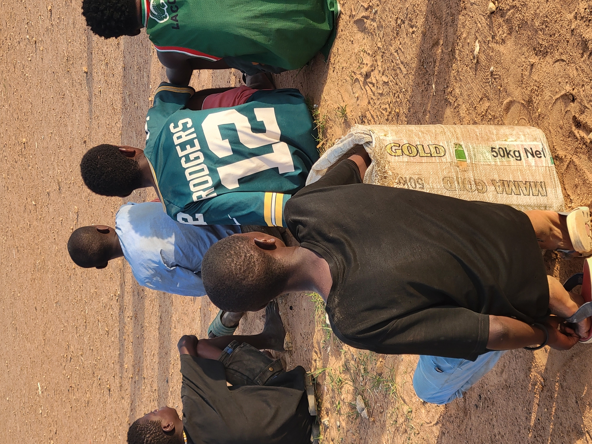 The image depicts a group of five young men outdoors, gathered around a sack labelled "GOLD 50kg Net" and "MAMA GOLD 50kg". They are located on bare, reddish-brown earth, possibly in a rural or undeveloped area. The men are dressed in casual clothing, including jerseys and t-shirts. One wears a jersey with "RODGERS 12" printed on it. The focus of the scene is the sack, around which the group is gathered. The setting appears to be sunny, based on the brightness and shadows. The location is stated as Ibo, Nigeria.