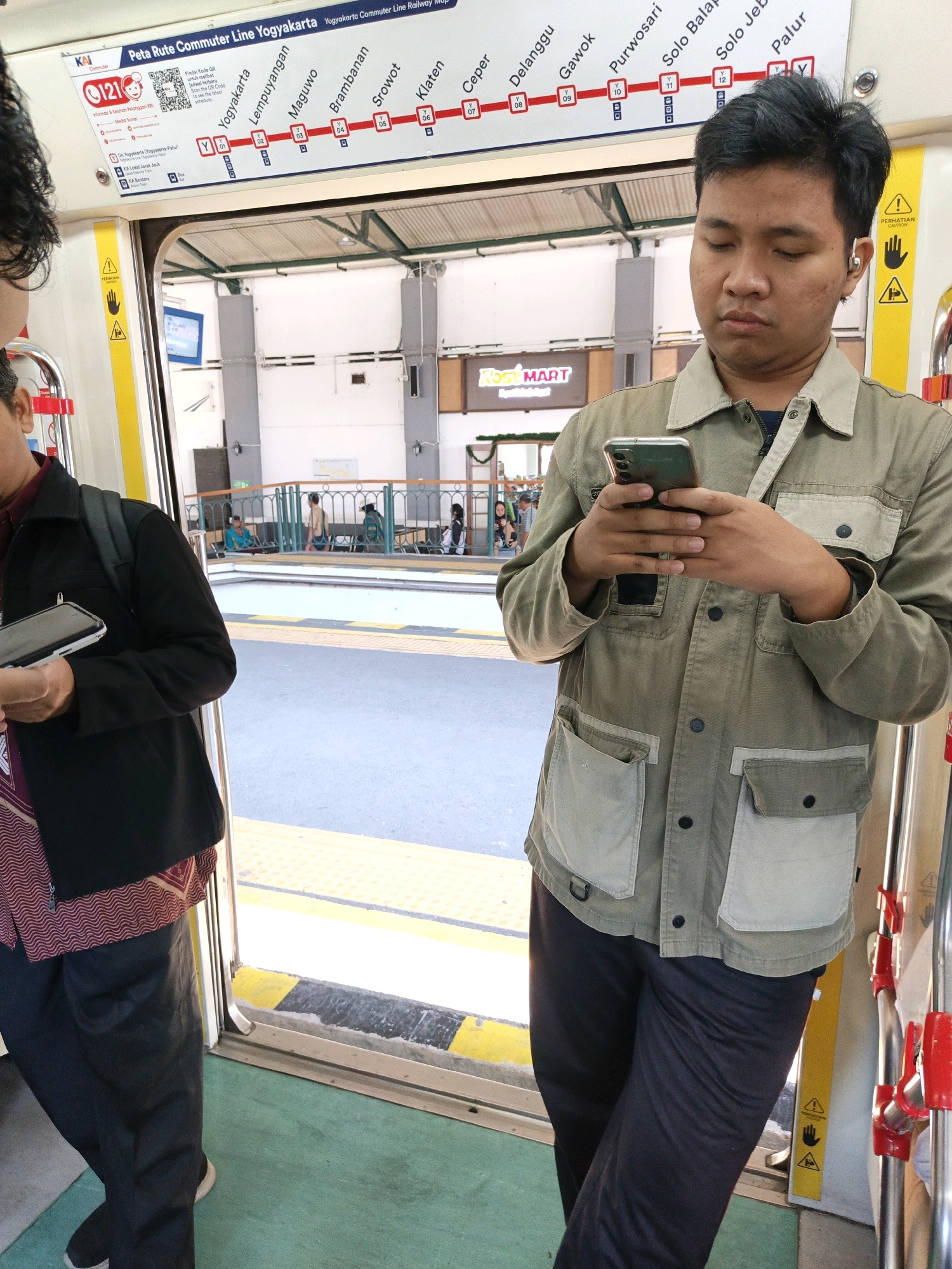 A young man stands inside a train car, holding a smartphone and looking down at it. He is wearing a light olive-green utility jacket with light beige pockets and dark navy pants. He has short, dark hair and is wearing a silver earbud in his right ear. Another person, only their upper body and legs are visible, stands to the left, also looking at a phone or device. They are wearing a black jacket over a patterned red and white shirt, and dark pants. The train car has yellow and black striped flooring and yellow handrails. Through the open door, a platform is visible with several other people waiting. In the background, a sign is visible indicating "Yogyakarta Commuter Line Railway Map" and names of various stations including "Yogyakarta", "Lempuyangan", "Maguwo", "Brambanan", "Srowot", "Klaten", "Ceper", "Delanggu", "Gawok", "Purwosari", "Solo Balapan", "Solo Jebres", and "Palur". The scene is likely a train station in or near Klaten, Indonesia.