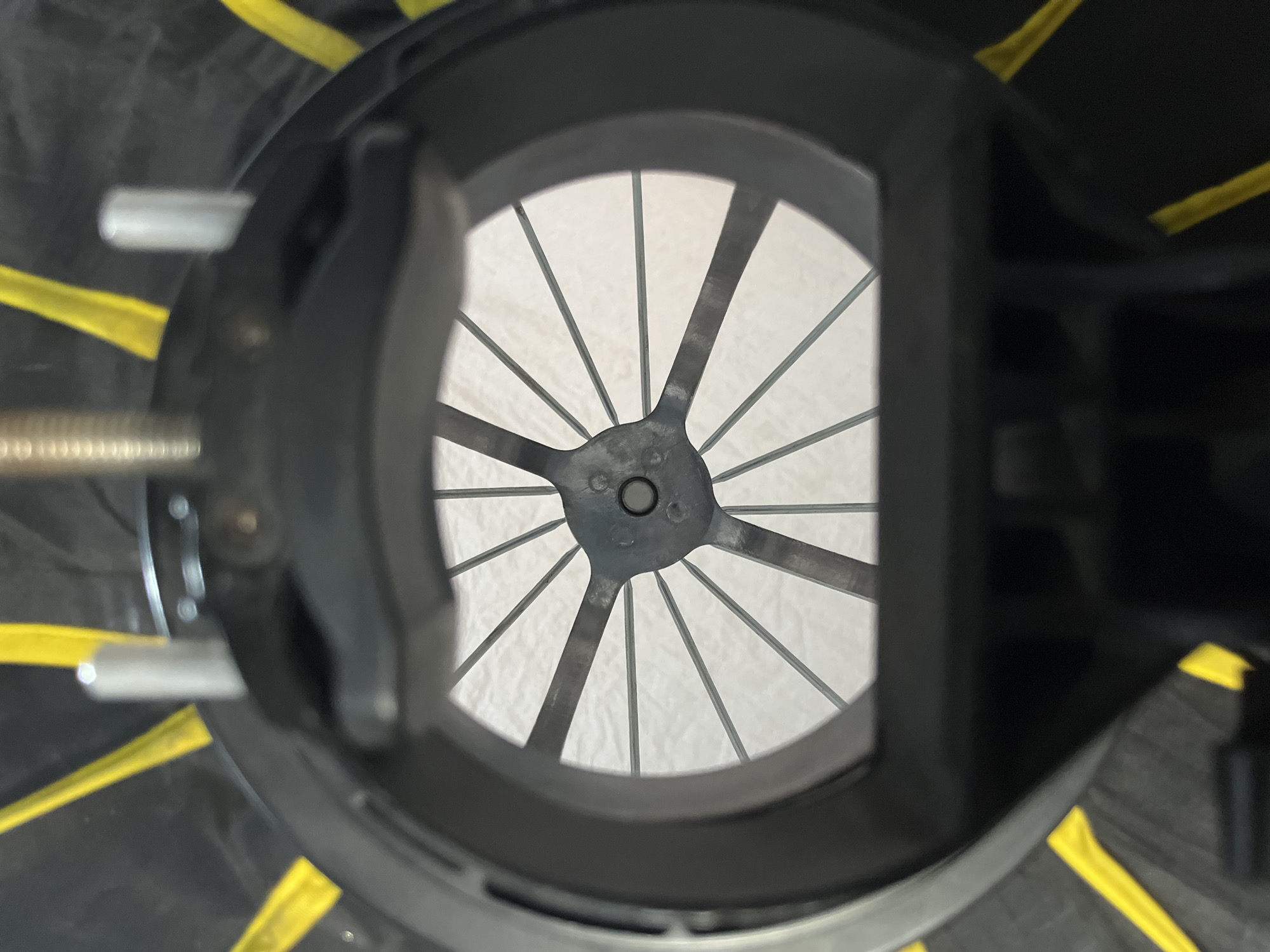 The image shows a close-up, through-the-lens view of a spoke-like structure. The perspective is from within a dark, cylindrical object, looking outwards. The spokes, approximately ten in number, radiate from a central hub. The hub is a dark, circular component with a small hole in its center and two visible fasteners or rivets. The spokes themselves are thin, metallic rods, possibly silver or gray in color, and they appear to be attached to a wider, darker ring towards the outer edge of the visible area.
Beyond the spoke structure, the background is a textured, off-white surface, possibly fabric or paper, with a slight crinkled appearance.
Framing the view from the front is a dark, rigid structure. This structure has a rectangular opening through which the spoke system is observed. On either side of this opening, and extending slightly beyond, are two vertical metallic rods, appearing silver and cylindrical. These rods seem to be part of the larger mechanism to which the observed object is attached.
The periphery of the image is filled with a black material, against which are angled, bright yellow, diagonal bands. These bands are curved and appear to be part of a fabric or flexible material, possibly a shield or cover.
There are no people, animals, or signs of life visible in the image. The environment appears to be indoors or a controlled setting, with the focus solely on the mechanical components. The lighting is direct, illuminating the spoke structure clearly. No specific location in Gura Top, Nigeria, can be identified from this image.