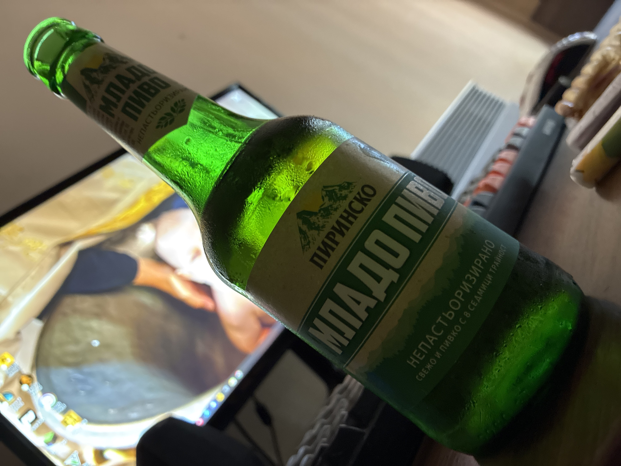 The image features a cold, green glass bottle of Bulgarian beer, prominently displayed on a surface, likely a desk. Condensation coats the bottle, indicating it is chilled. The label, written in Cyrillic, clearly shows "МЛАДО ПИВО" (Mlado Pivo), which translates to "Young Beer." Above this, "ПИРИНСКО" (Pirinsko) is visible, suggesting the brand name, likely referencing the Pirin Mountains. Additional text on the label includes "НЕПАСТЬОРИЗИРАНО" (Unpasteurized) and "СВЕЖО И ПИВКО С 8 СЕДМИЦИ ТРАЙНОСТ" (Fresh and drinkable with 8 weeks shelf life).
In the blurred background, a computer monitor is visible, displaying what appears to be a classical painting or digital artwork featuring warm hues and a human form. To the right of the bottle, a keyboard with distinct orange and white keys, along with other desk accessories, can be seen. The overall setting is indoors, with ambient lighting creating a relaxed atmosphere. No people are visible in the photograph. This scene, featuring a Bulgarian beer, is consistent with the provided location context of Varna, Bulgaria, depicting a casual moment at a workstation.