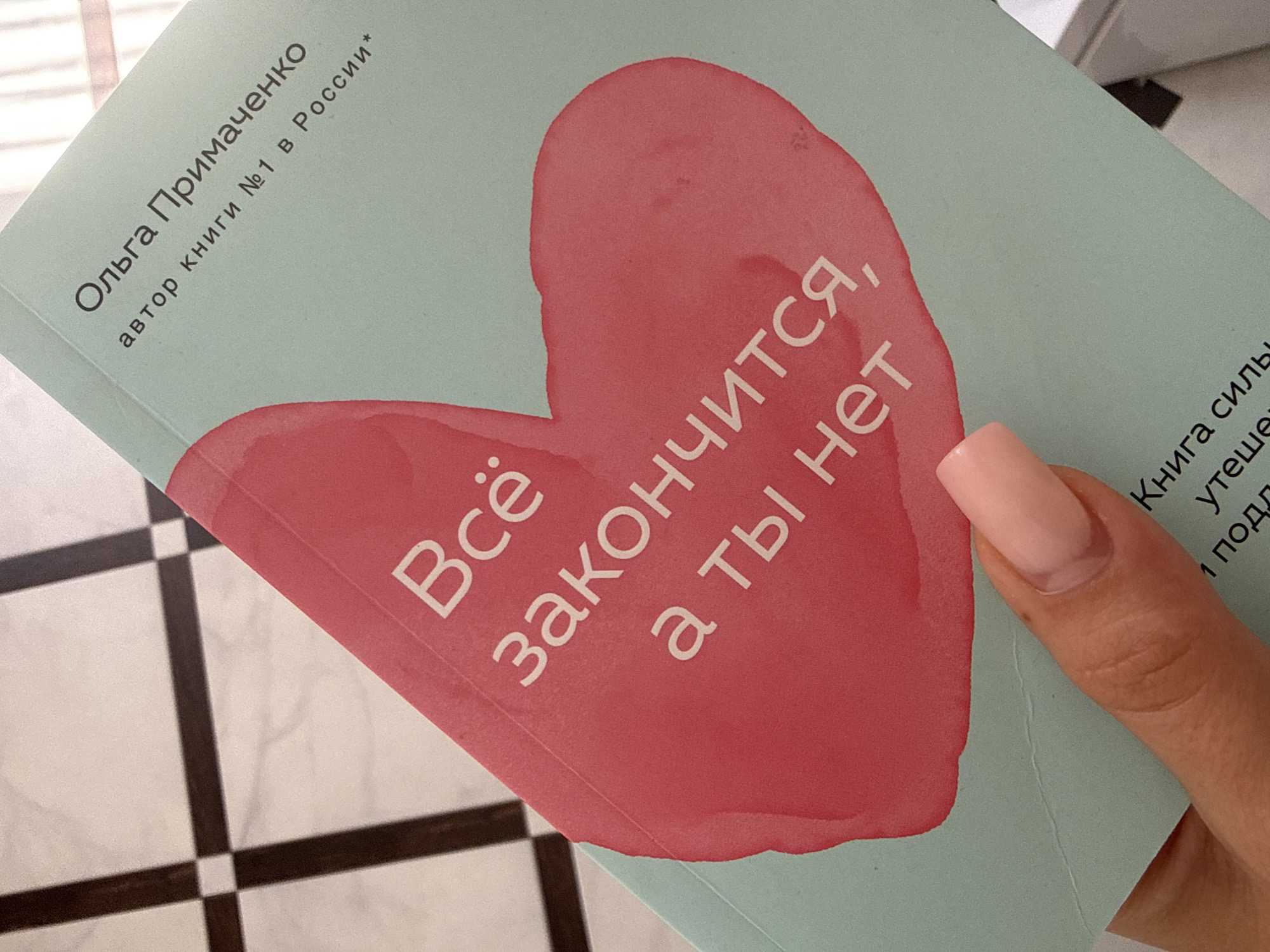 In this close-up, top-down shot, a light teal book with a pink heart illustration is held by a person's hand, revealing a light pink acrylic nail. The book is angled diagonally. The title on the heart reads in white Cyrillic text, "Всё закончится, а ты нет". Above the heart, in smaller white Cyrillic text, is "Ольга Примаченко" and "автор книги №1 в России*". At the bottom of the book, partially visible text in white Cyrillic reads "Книга силы" and "Утеши". The background is a black and white tiled floor, possibly marble or ceramic, with dark brown grout lines forming a grid pattern. The scene appears to be indoors, possibly in a home or a public space, and does not provide any specific indicators of location within Chișinău, Moldova.