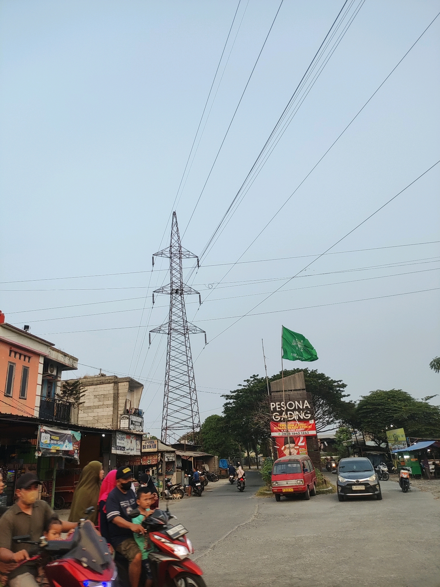 The image depicts a street scene in Wanajaya, Indonesia, under an overcast sky. Dominating the background is a large electricity pylon. In the middle ground, several buildings line the street, including one with an orange facade. Various small shops with awnings are visible. Traffic is present, with several motorcycles carrying passengers, including families, in the foreground. A red van and a dark-colored car are stopped at the intersection. A green flag with text and a crest is flying from a flagpole. The overall environment suggests a bustling, urban setting.