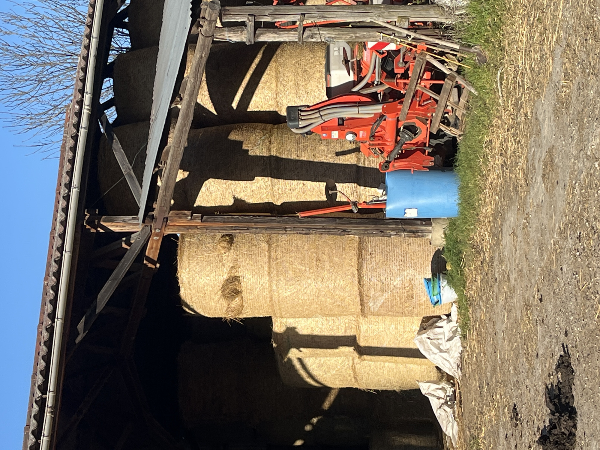 The image depicts a barn filled with stacked hay bales in Chambœuf, France. An orange agricultural machine with a blue water tank is parked in front of the hay bales. The barn is made of wood and corrugated metal. To the right of the barn, there is a patch of grass and dirt. Some white tarps are covering the lower hay bales.