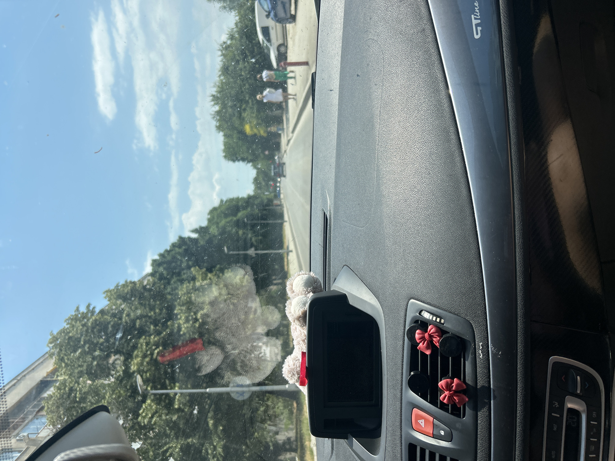 The image is taken from inside a car, looking out through the windshield. The car is driving on a road in the city of Plovdiv, Bulgaria. The road is lined with trees on both sides, and there are buildings visible in the distance. The sky is blue with some clouds. On the dashboard of the car, there is a car air freshener in the shape of a teddy bear with a red bow, and two red bows on the air vents. There are also two people walking on the sidewalk. One person is wearing a white dress, and the other is wearing green pants. There are several cars on the road, and a white van is parked on the side of the road. The overall impression is that of a bright, sunny day in a city.