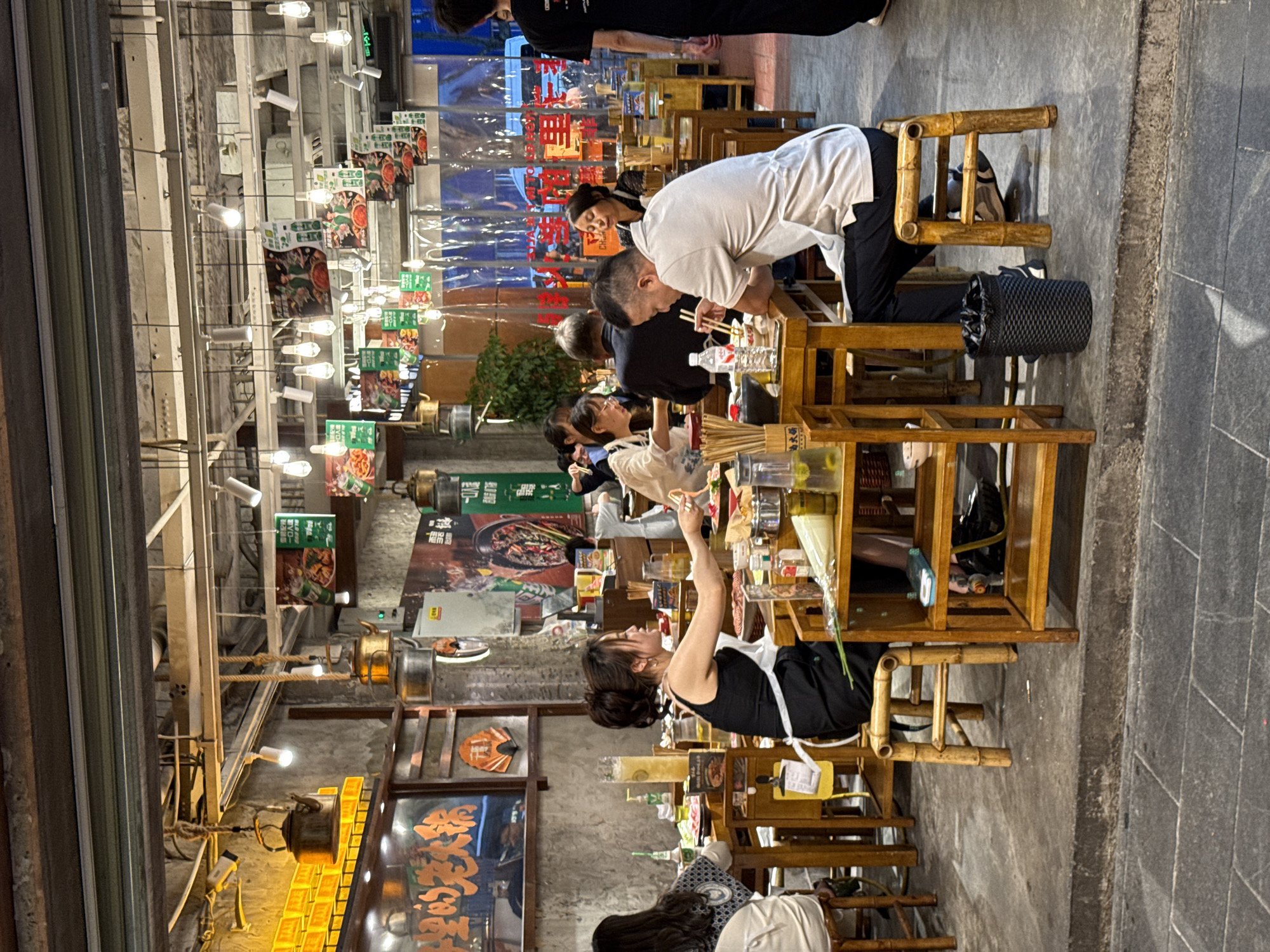 The image shows an outdoor dining area in the Jinjiang District, China. Diners are seated at wooden tables and chairs on a gray stone-paved street. A server in a black tank top and white apron is interacting with a table. Above the diners are hanging lights and advertisements for food. Plastic sheeting is visible in the background. Traditional teapots and other decorative elements hang from the building.