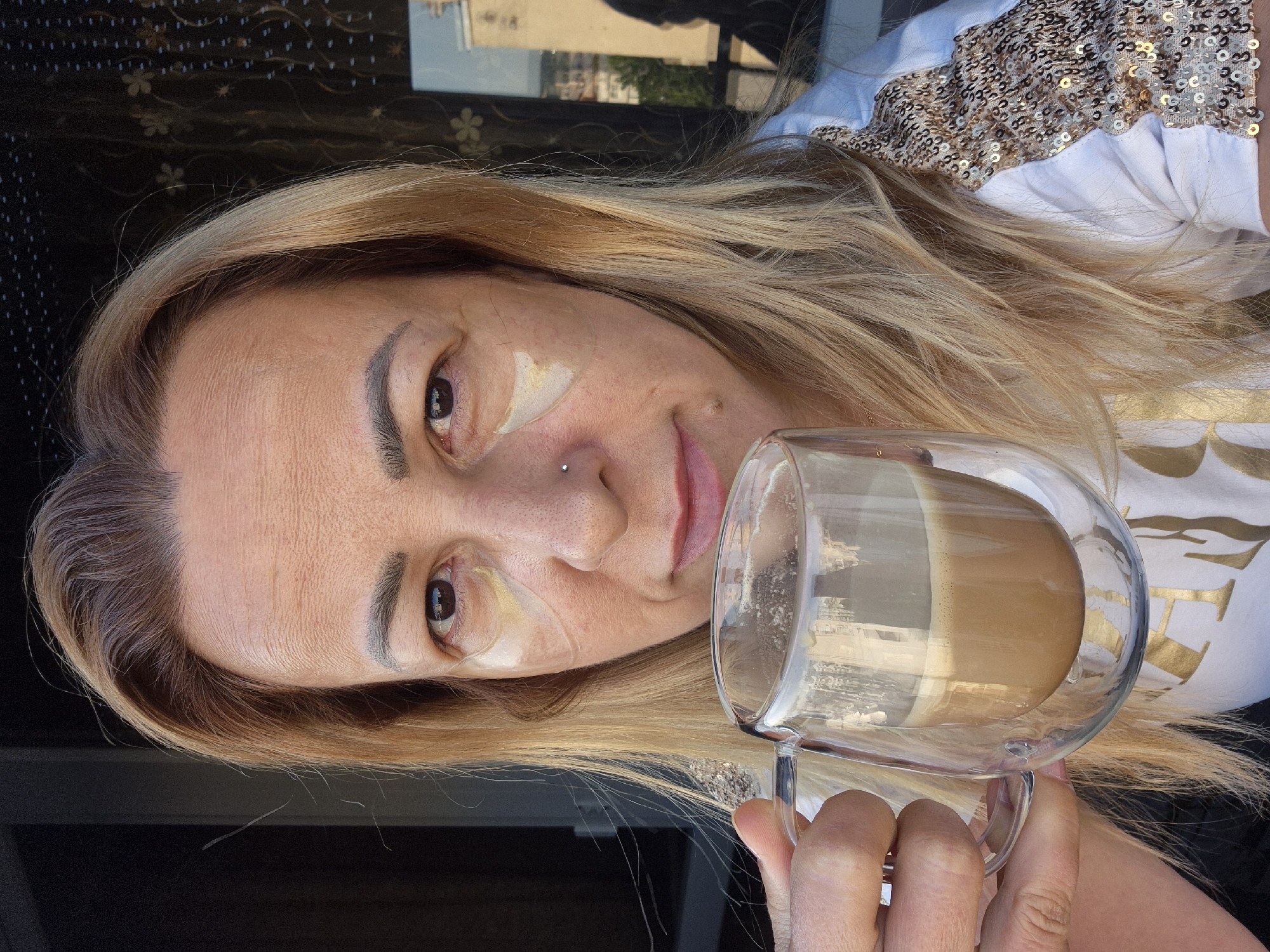 A medium close-up shot shows a woman with blonde hair drinking coffee from a clear double-walled glass mug. She is looking at the camera and has eye patches under her eyes. She is wearing a white t-shirt with gold lettering and sequined sleeves. The background is out of focus, showing a window with a patterned curtain and a reflection of buildings.