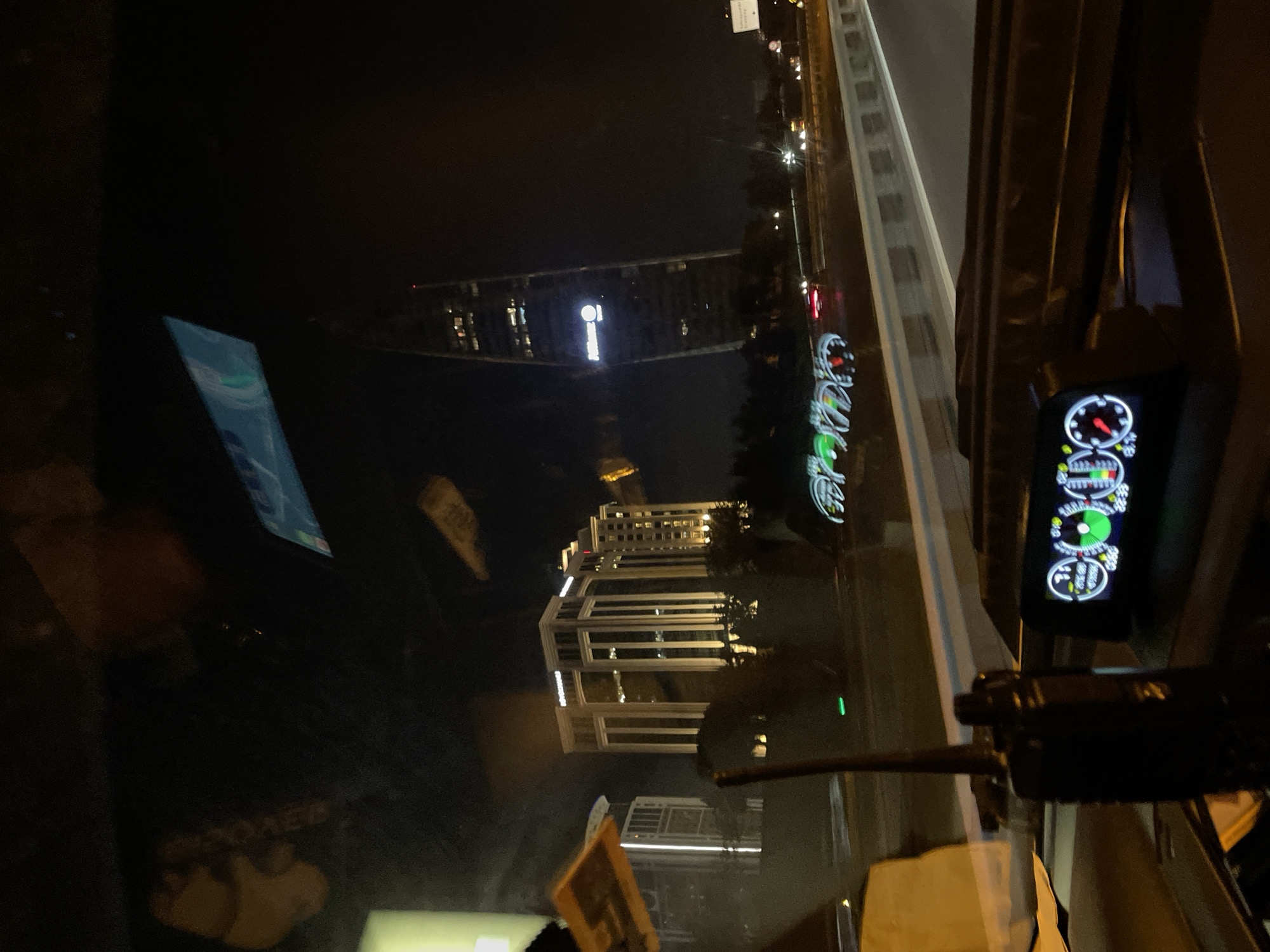 The image shows a view from inside a vehicle at night, looking out at a city skyline. In the foreground, a digital gauge cluster with a circular display is visible on the dashboard. To the left of the cluster, a black walkie-talkie with an antenna is mounted. The windshield reflects some lights and objects, including a blue and white sign with text and a green horizontal line.
Outside the vehicle, several illuminated buildings are visible. A large, modern office building with a distinctive curved shape and a glowing logo that appears to read "Allianz" is prominent on the right side. To the left, a rectangular building with multiple floors and numerous illuminated windows stands out. The buildings are set against a dark night sky, with some trees and a highway visible in the mid-ground. The highway has a white barrier and some distant car lights.
The scene is likely the city of Ataşehir, Turkey, as indicated by the presence of the Allianz Tower, a well-known landmark in that district of Istanbul. The time appears to be nighttime, given the darkness and the illumination of the city.
