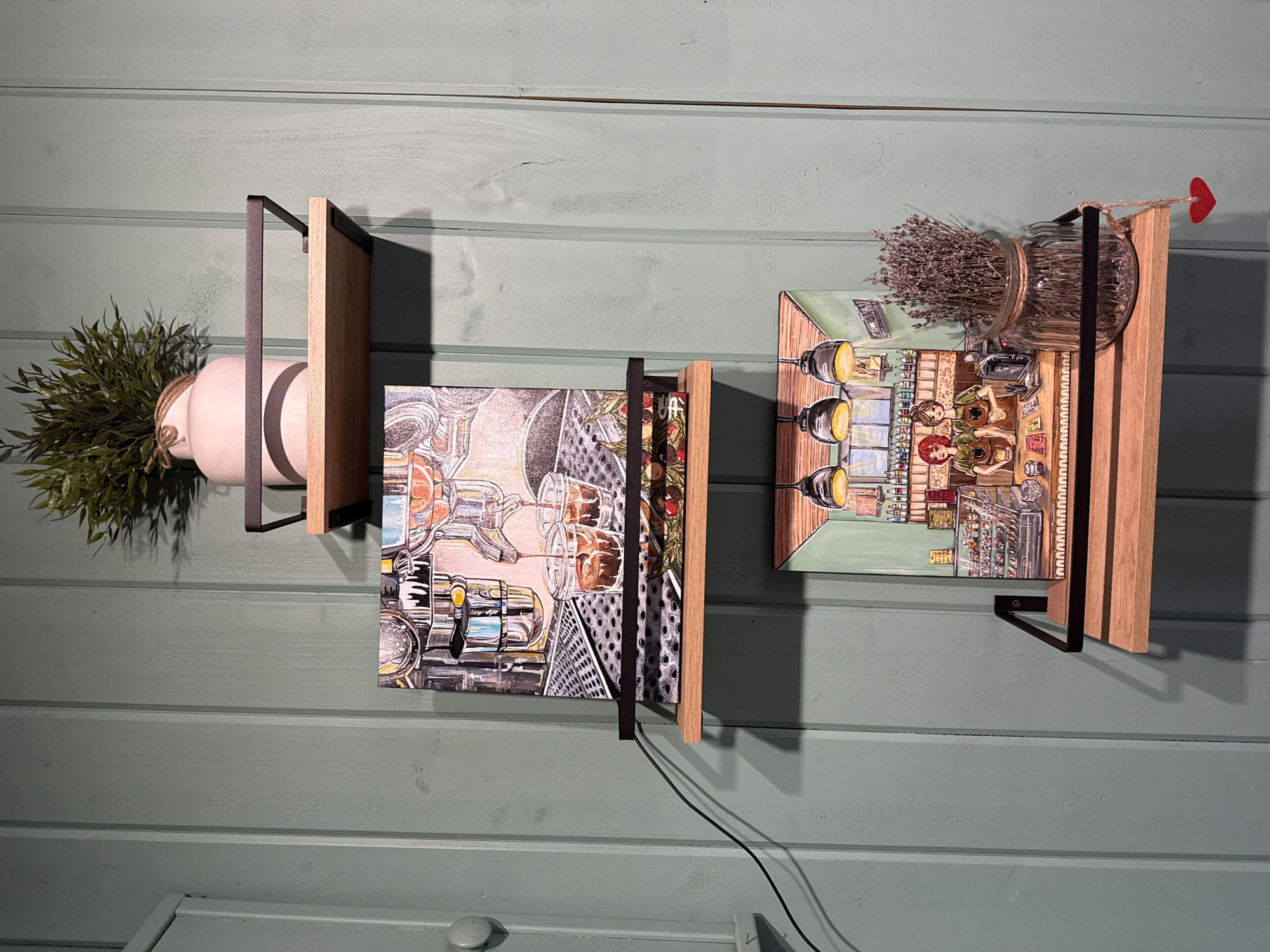 A view of two wall-mounted shelves with art and decor. The wall is painted light green with visible horizontal brushstrokes. The shelves are wooden with black metal brackets. The left shelf holds a round white pot with green leafy plants spilling out. The right shelf displays an art piece depicting what appears to be an interior scene with figures, alongside a glass jar filled with dried lavender, a small red heart-shaped ornament hanging from the jar with twine. A black cord runs down from behind the artwork on the left shelf.