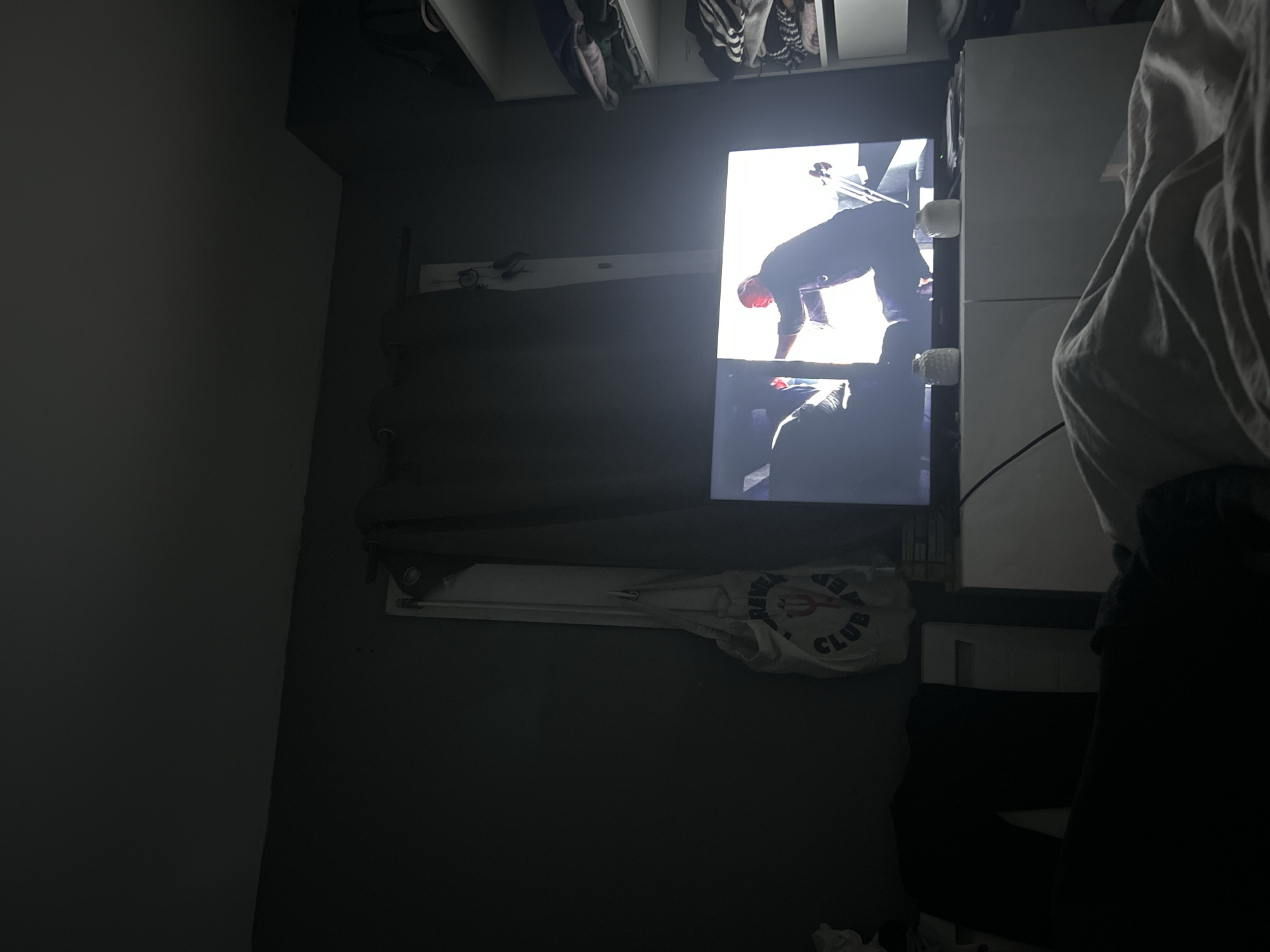The image depicts an indoor scene featuring a television screen mounted on a wall, displaying a video. In the video, a person is shown in a forward bend pose, with their hands likely touching the ground. To the left of the television screen, a white bag with the words "REVEALER CLUB" is visible. A partially visible wardrobe is positioned above the TV, while a person stands in front of the TV, partially obscuring it. The location is potentially Lannoy, France, based on the text on the bag.