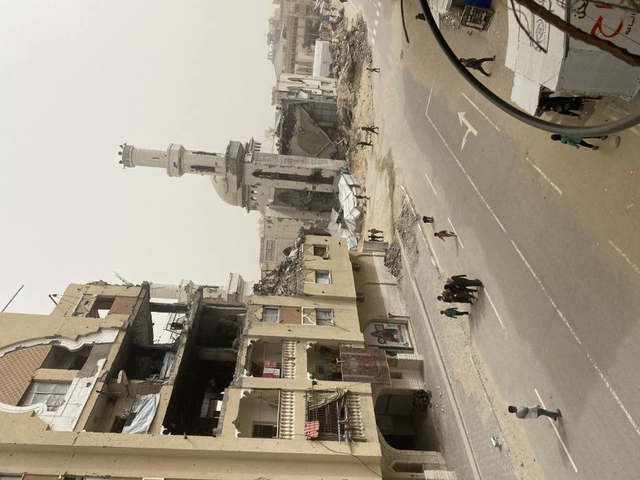 This image depicts a street scene in Khan Yunis, Palestinian Territory, characterized by extensive structural damage under an overcast sky. In the foreground-left, a multi-story building shows severe damage, with large sections of its facade missing, exposing internal floors, collapsed elements, and visible rebar; laundry hangs from balconies on multiple levels. To the center-right, a mosque structure stands partially destroyed, featuring a largely intact minaret but with significant damage to its dome and main walls, surrounded by substantial piles of rubble. The paved street below features white lane markings, including a turn arrow, and is traversed by approximately fifteen individuals, comprising both adults and children, some walking in small groups and others singly. On the right side of the street, amidst the debris, several temporary shelters constructed from white tarpaulins are visible. Additional damaged structures and scattered debris extend into the background.