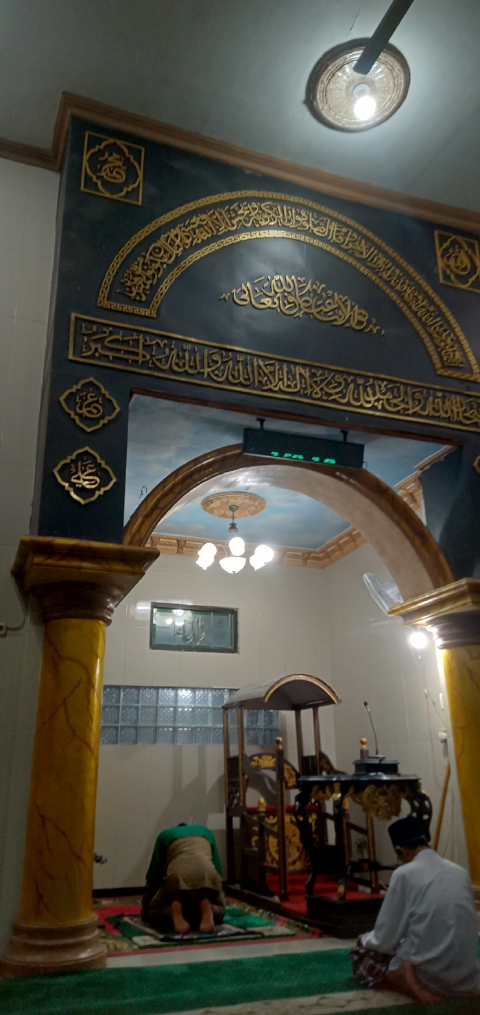 The image displays the interior of a mosque in Perak, Indonesia. Two male individuals are engaged in Islamic prayer on prayer mats. In the mid-ground to the left, one individual is in the sujud (prostration) position, wearing a green top and light brown lower garment. In the foreground to the right, another individual sits facing the same direction, dressed in a white long-sleeved shirt and a patterned sarong.
The central background features a dark wall adorned with intricate gold Arabic calligraphy arranged in an arch, signifying the qibla direction. Above this arch, a digital display board with green characters is mounted. Two prominent yellow-marbled columns flank the prayer area. Behind these columns, an ornate brown archway opens to another section of the room with a light blue, cloud-painted ceiling and a multi-bulb chandelier. To the right of the praying individuals, a wooden minbar with golden decorative elements and a dark wooden lectern with a microphone are positioned. On the left wall, a rectangular window with patterned glass and a section of glass block wall are visible. The ceiling in the foreground includes a circular light fixture and part of a ceiling fan. The scene is brightly illuminated by artificial light.