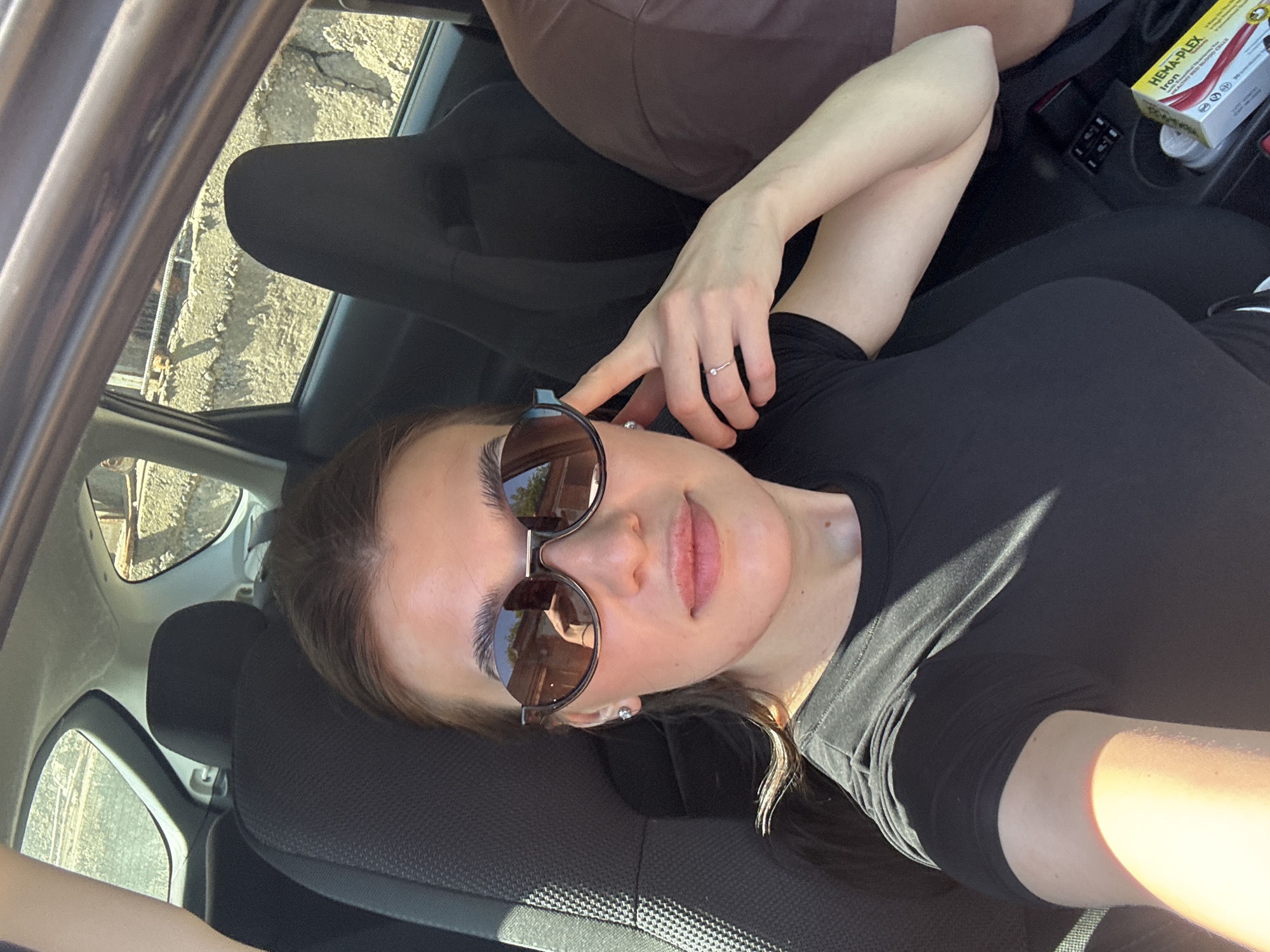 A female individual is observed seated within the interior of a vehicle, in what is identified as Chaikovsky, Russia. The perspective suggests a selfie taken from a front seat position. The individual has brown hair, fair skin, and full lips, and is wearing dark, semi-rimless sunglasses with dark frames. A small stud earring is visible on her left ear, and a thin, silver-toned ring with a single small stone is on her right ring finger. She is dressed in a black short-sleeved top. Her right hand is slightly raised, with fingers touching the right temple area near the sunglasses. Strong sunlight enters the vehicle, creating highlights on her skin and clothing. Through the vehicle's right rear window, an exterior environment is visible, comprising a textured concrete wall or embankment, horizontal metallic rebar structures above it, and distant light-colored buildings. Reflections in the sunglasses show parts of the car's interior. On the lower right, near the center console, a yellow and red box labeled "HEMA-PLEX Iron" and a white container are present.