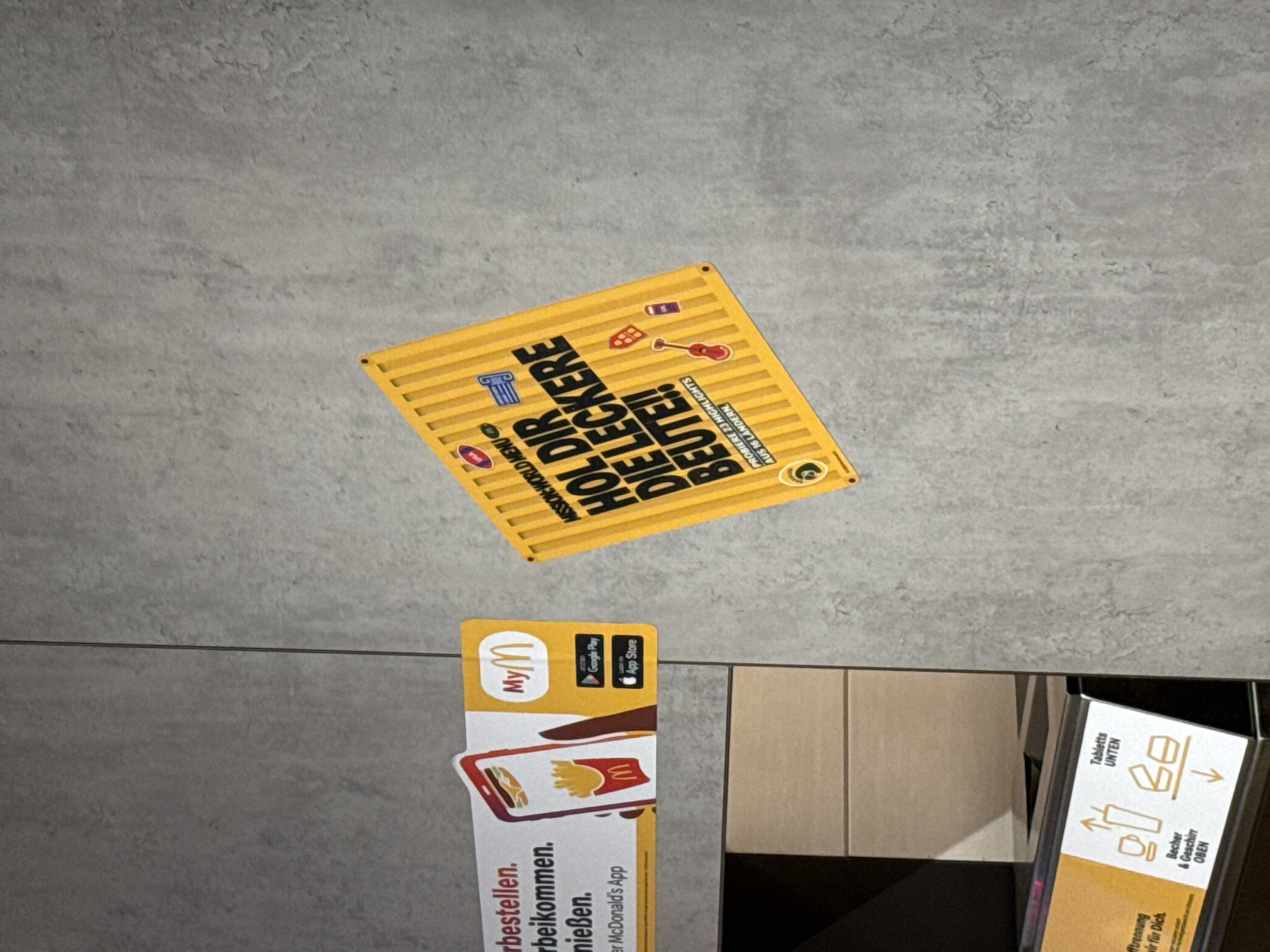The image shows a section of a concrete wall with various stickers and signage. On the left, a white and orange sticker advertises the "MyMcDonald's App" with a depiction of a phone displaying a burger and fries. Below this sticker, a portion of a shelf unit is visible. To the right of the MyMcDonald's sticker, a bright yellow square sticker with bold black text reads "HOL DIR DIE LECKERE BEUTE!!". This sticker features various smaller icons and the text "PROBIERE 23 HIGHLIGHTS AUS 16 LÄNDERN". The concrete wall has a textured, grey appearance. In the lower left corner, another sign with orange and white elements displays icons related to trash disposal, indicating "Becher & Geschirr OBEN" (cups and cutlery above) and "Tabletts UNTEN" (trays below). The presence of the McDonald's branding indicates this scene is likely within a McDonald's restaurant, possibly in Saalfeld/Saale, Germany, given the German language on the signage. There are no people visible in the image.