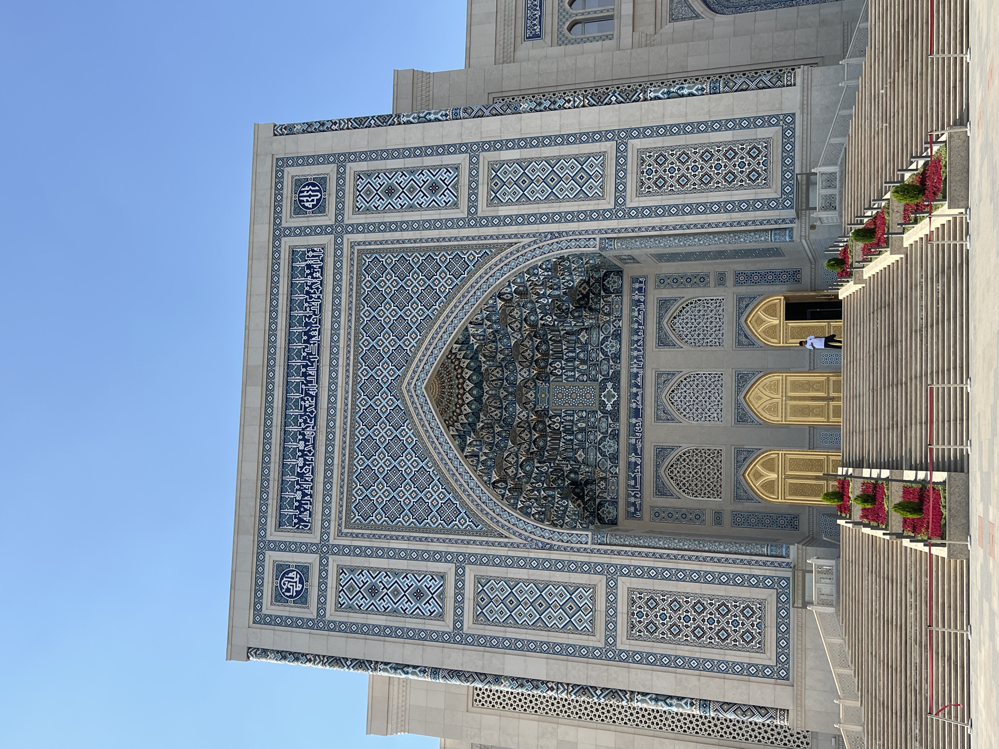The image shows the exterior of the Hazrat Imam Mosque, also known as the Hast Imam or Khast-Imam complex, in Tashkent, Uzbekistan. The mosque features a grand entrance with intricate blue and white geometric tilework and Arabic calligraphy. A wide staircase leads up to the golden doors of the mosque. Red stanchions and ropes guide visitors up the stairs, and flower beds with red and green foliage flank the central section of the staircase. A single person, dressed in a white shirt and dark pants, is visible standing near the open entrance. The sky above is clear and blue, indicating a sunny day.