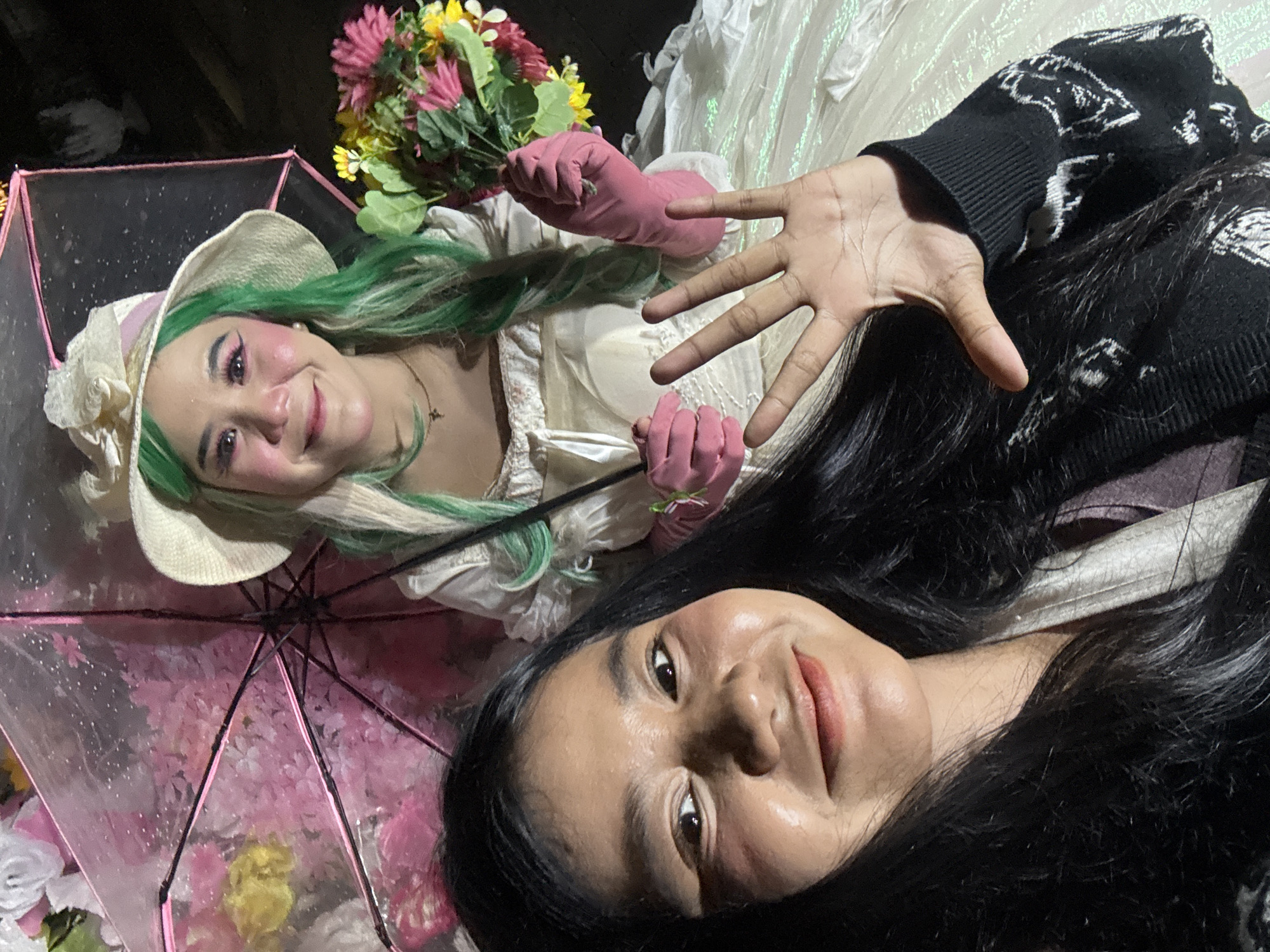 Two individuals are posing for a selfie outdoors at night. The person in the foreground is closer to the camera and is looking directly at it with a smile. They have long, dark hair and are wearing a black sweater with a white pattern. Their right hand is raised, showing all five fingers extended towards the camera. The second individual is positioned behind the first and to the right. They are wearing a white hat with a pink and white ribbon, a white dress, and bright pink gloves. They also have bright green hair with blonde streaks. They are holding a bouquet of colorful flowers in their right hand. Behind them, a clear umbrella decorated with pink flowers is visible, with some of the flowers appearing to be attached to the umbrella's spokes. The background is dark, suggesting it is nighttime. The image does not contain any specific visual cues to determine the exact location within the city of West Jakarta, Indonesia.