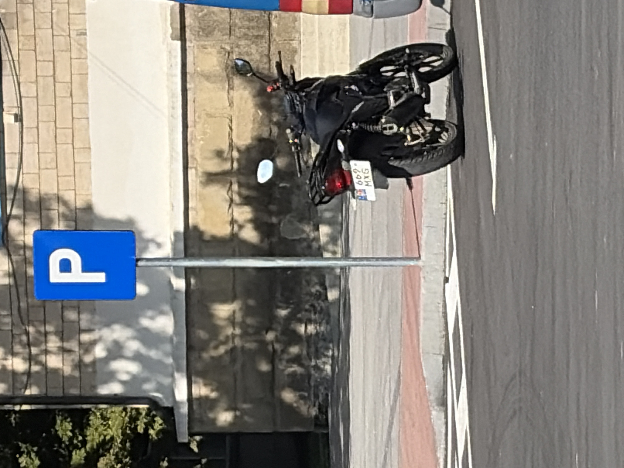 A black motorcycle is parked on a paved surface adjacent to a sidewalk in Criuleni, Moldova. The motorcycle is facing away from the camera and has a license plate that reads "669 MXG". A blue parking sign with a white "P" is visible to the left of the motorcycle. In the background, a building with a stone facade is visible, with sunlight casting shadows on its surface. The scene appears to be an outdoor urban environment.