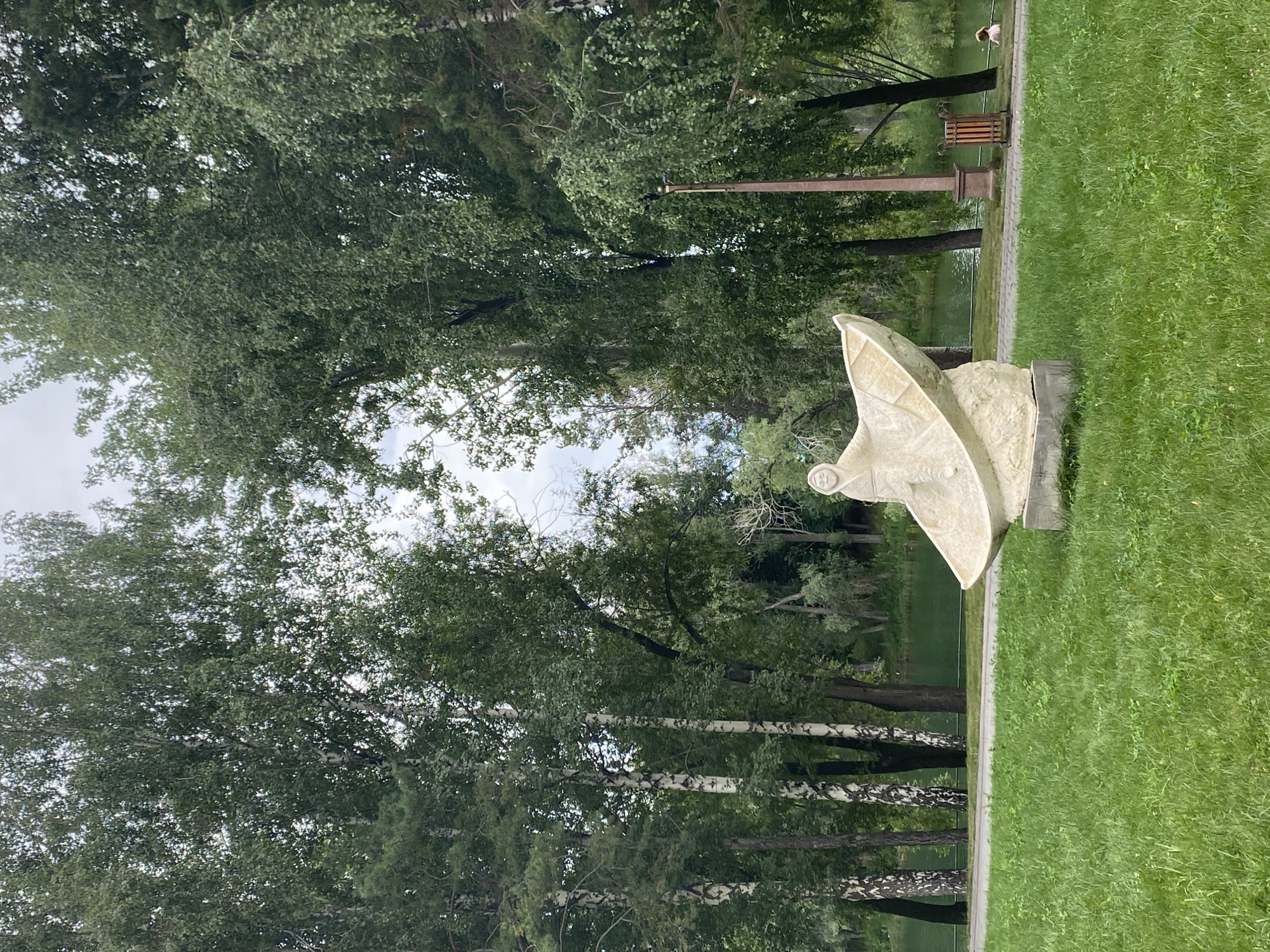 The image captures a serene outdoor scene in Cholpon-Ata, Kyrgyzstan. In the foreground, a light-colored stone sculpture depicts a figure seated in a boat-like vessel, positioned on a manicured green lawn. Behind the sculpture, a paved pathway runs parallel to a body of water, likely Lake Issyk-Kul, with lush green trees lining the far bank. The trees vary in type, including birch trees with distinctive white bark and other deciduous trees with dense foliage. The sky is overcast, suggesting a cloudy day. Further in the distance, a person is visible sitting near the water. A lamppost and a trash receptacle are situated on the pathway, adding functional elements to the park-like setting. The overall atmosphere is tranquil and natural.