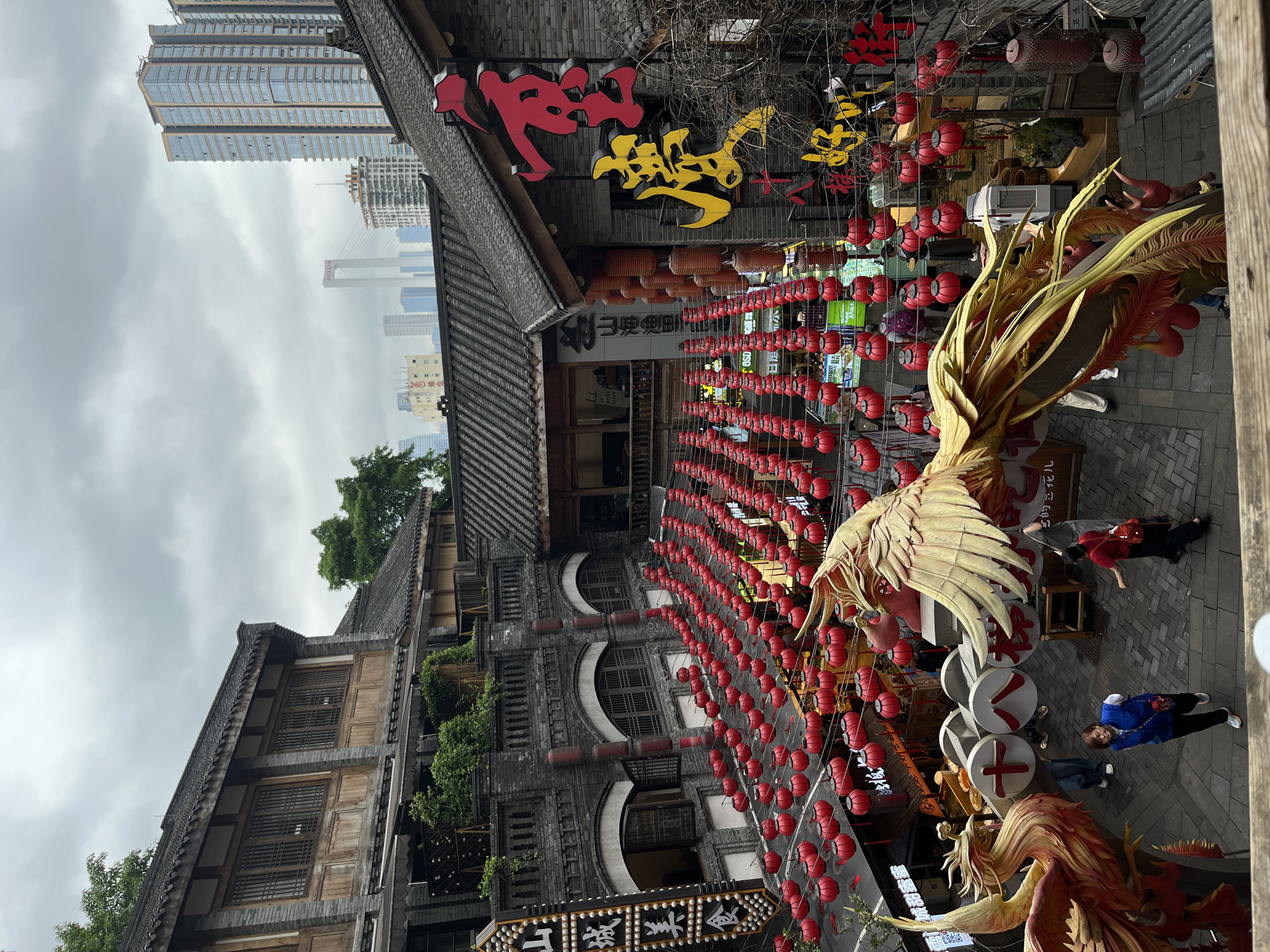 The image captures a street scene in Yuzhong District, Chongqing, China. The street is adorned with numerous strings of red lanterns. There is a large golden phoenix statue prominently featured on the right side of the frame. People are seen walking around the statue. The architecture consists of traditional Chinese-style buildings, with tiled roofs and wooden facades. In the background, modern skyscrapers are visible, creating a contrast between old and new architecture. Chinese characters are displayed on signs above the buildings. The weather appears to be overcast, with a cloudy sky.