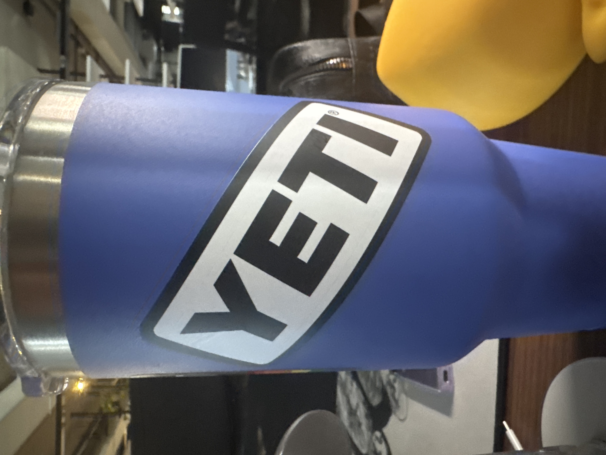A close-up, eye-level shot focuses on a blue YETI tumbler with a white and black YETI logo. The tumbler is the central subject, positioned in the foreground. The background is blurred and appears to be an indoor setting, possibly an office or workspace, with hints of office equipment and furniture. The lighting suggests artificial illumination. The scene is devoid of people and action. The exact location in Kyiv, Ukraine, cannot be determined from the visual information provided.