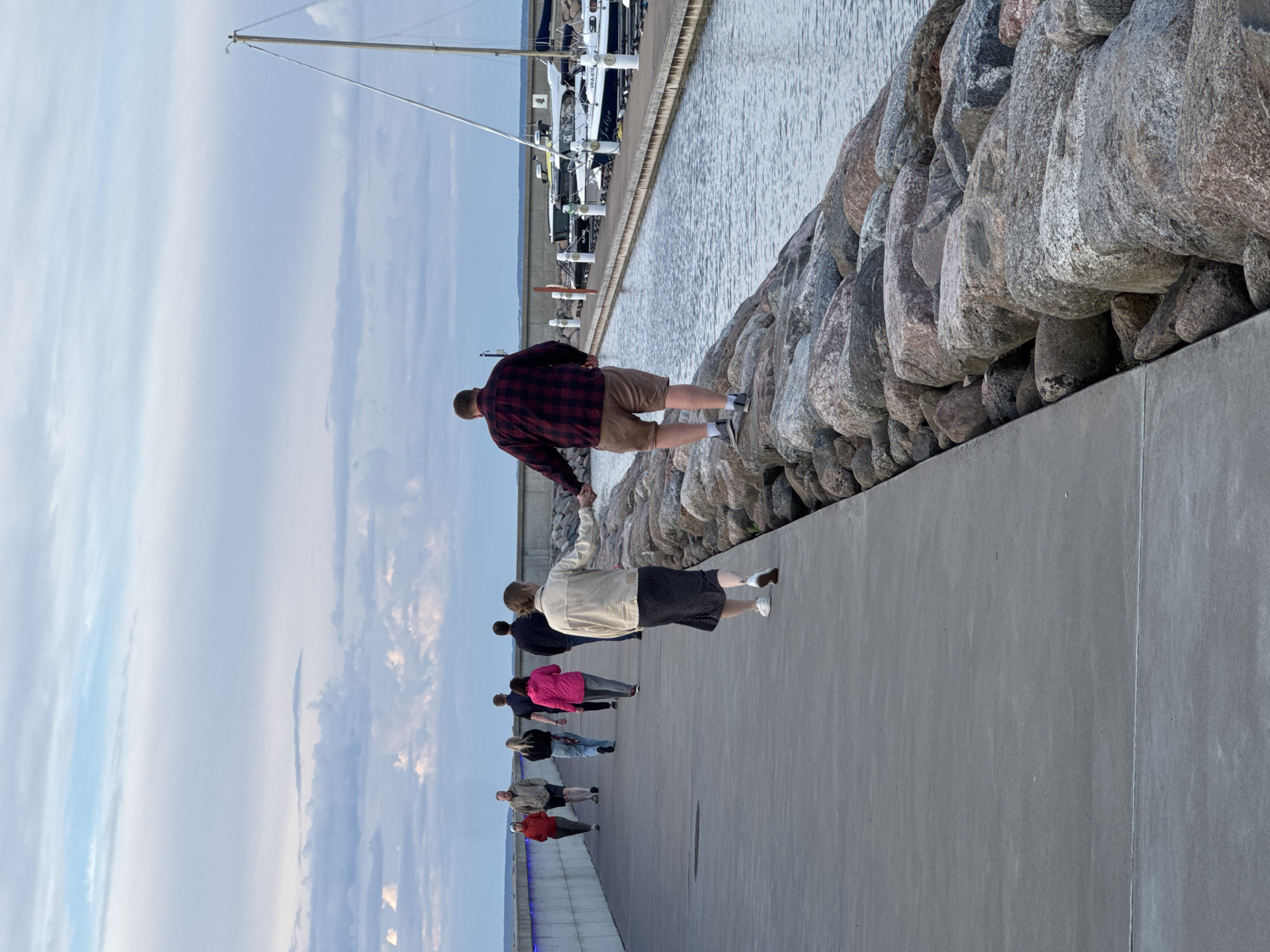 The image shows a pier with a concrete walkway and a rock embankment. Several people are walking on the pier. In the foreground, a man and a woman are walking hand-in-hand, with the man in a red and black plaid shirt and khaki shorts, and the woman in a beige jacket and a black skirt with white polka dots. Behind them, a group of people are walking in the same direction. The water on the right side of the pier has several boats docked. The sky is cloudy, with hints of blue. This scene appears to be at a marina in Tallinn, Estonia.