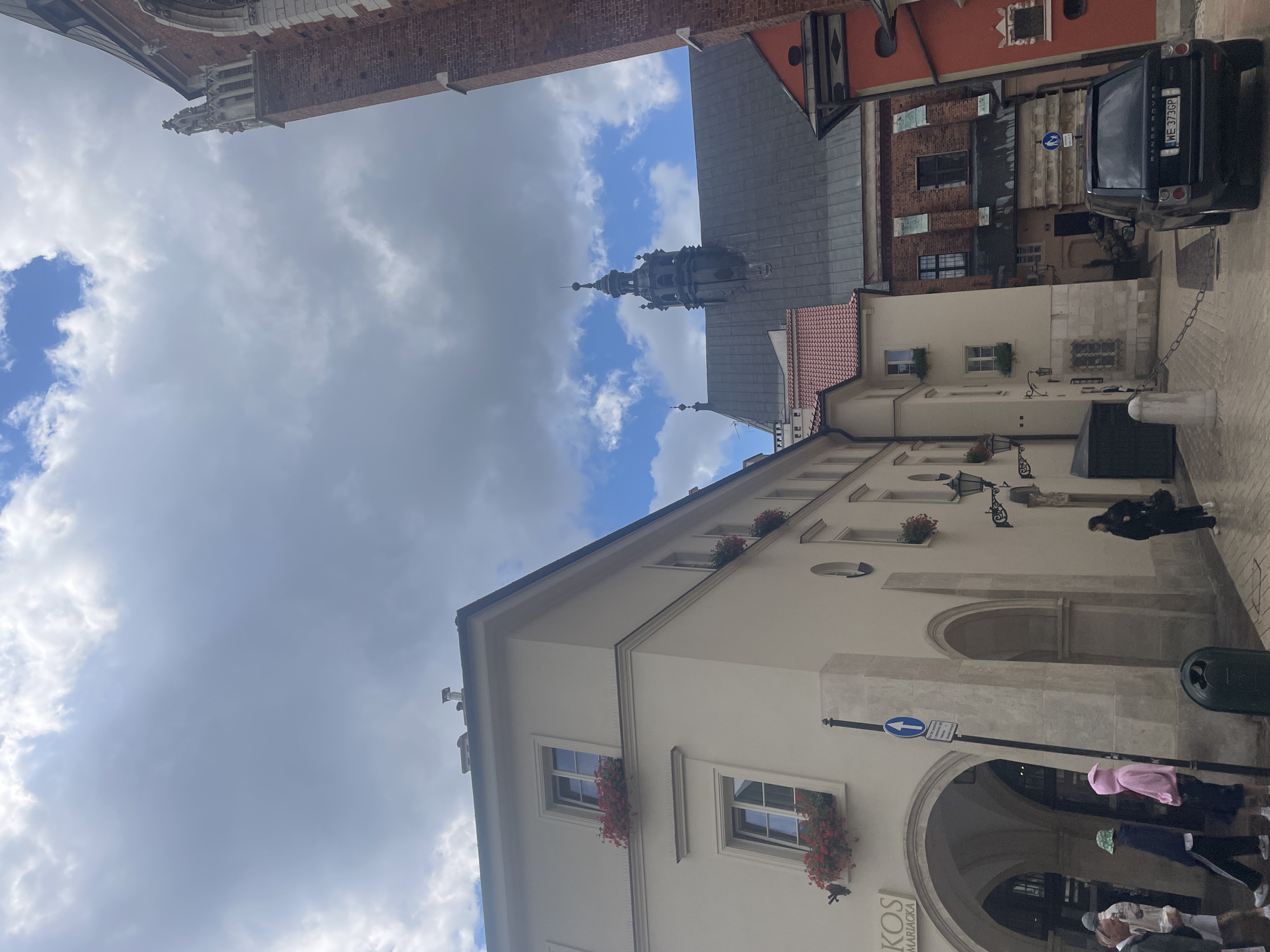 The image depicts a street scene in Krakow, Poland, likely in the Old Town. In the foreground, a black Range Rover with the license plate "WE 373GP" is parked on a cobblestone street. To the left of the vehicle, an archway leads into a building with text above it that is partially obscured but appears to include "KOS MARIACKA". Several people are visible near the archway, including a person in a pink hoodie and another wearing a green bucket hat.
Dominating the background are architectural elements of old buildings. A prominent brick tower with gothic detailing rises on the right side of the frame. To the left of this tower, a dark-tiled roof with a spire-like structure is visible, suggesting a church or another significant historical building. Further to the left, a cream-colored building with multiple windows, some adorned with red flowers, is prominent. The sky above is partly cloudy, with patches of blue sky visible amongst the white and grey clouds. The overall impression is of a historic urban environment on a slightly overcast day.