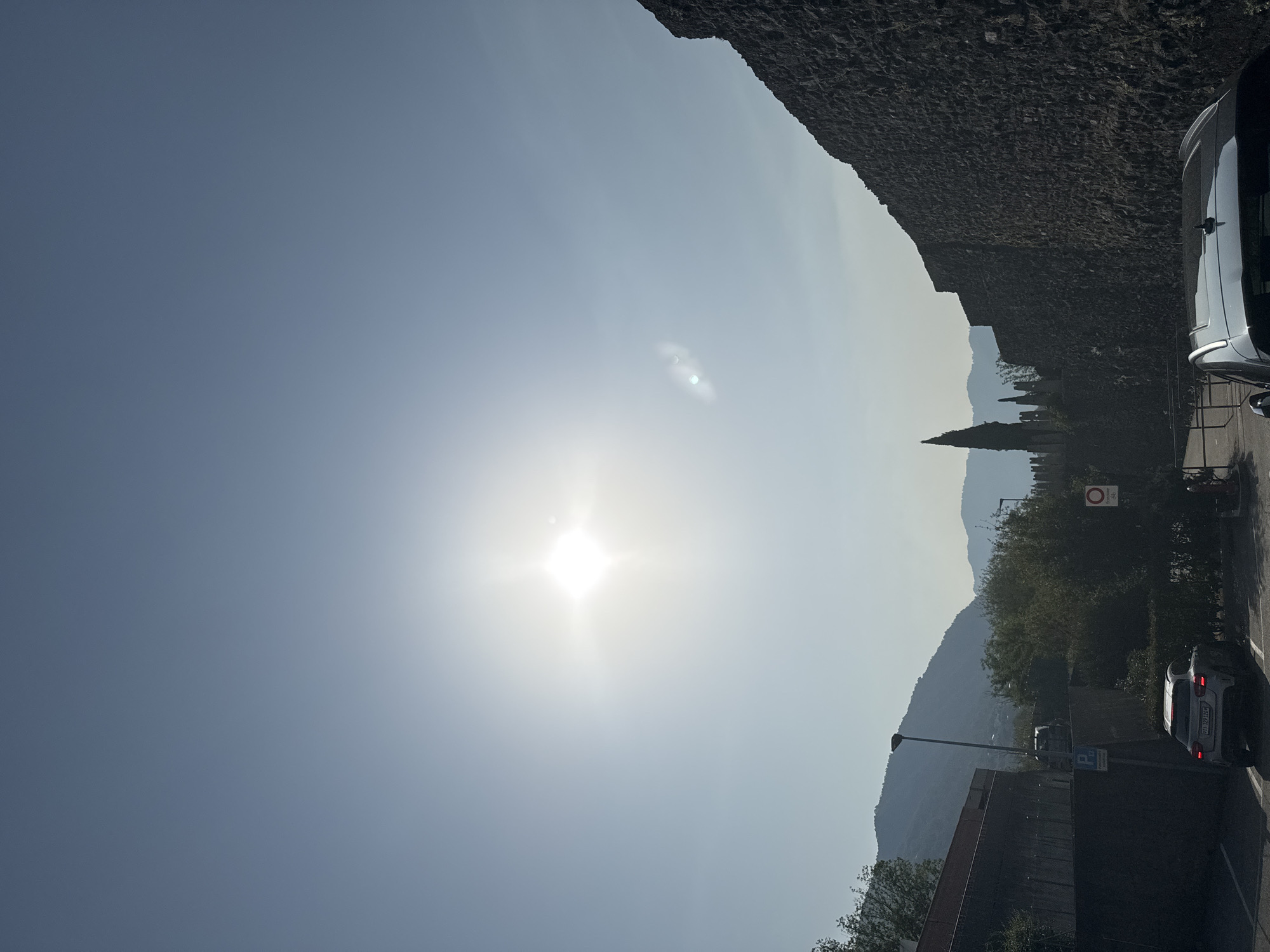 The image depicts a bright, sunny day with the sun prominently visible in the upper center of the frame, casting a strong glare. The sky is a clear, pale blue. In the foreground, a dark, textured stone wall extends from the right side of the image diagonally upwards. To the left, a grey sedan is parked in a paved area, and further back, a white SUV is visible. Behind the vehicles, there is a dense line of green trees and bushes. A tall, slender lamppost stands near the center. In the background, rolling hills or mountains are silhouetted against the hazy sky. A thin, spire-like tree, possibly a cypress, is visible rising above the wall on the left side. The image appears to be taken from a street or parking area in Balerna, Switzerland, with a view towards the surrounding landscape.