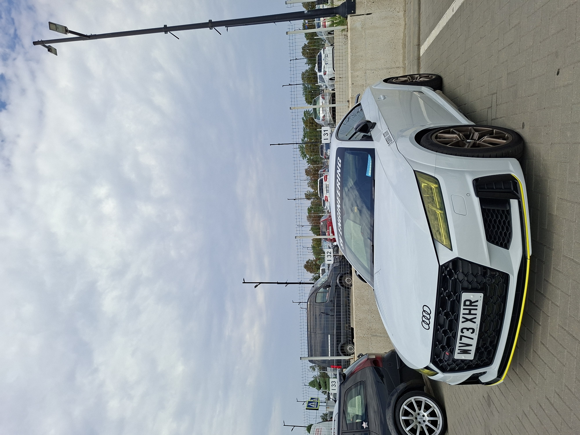 A white Audi TT RS is parked in a lot in Stăuceni, Moldova. The car is facing the viewer and is angled slightly to the right. It has a yellow stripe on the front bumper and yellow headlights. The license plate is WV73 XHR. A black Volvo wagon is parked to the left of the Audi. In the background, there is a chain-link fence, a concrete barrier, and several other cars parked. The sky is cloudy.