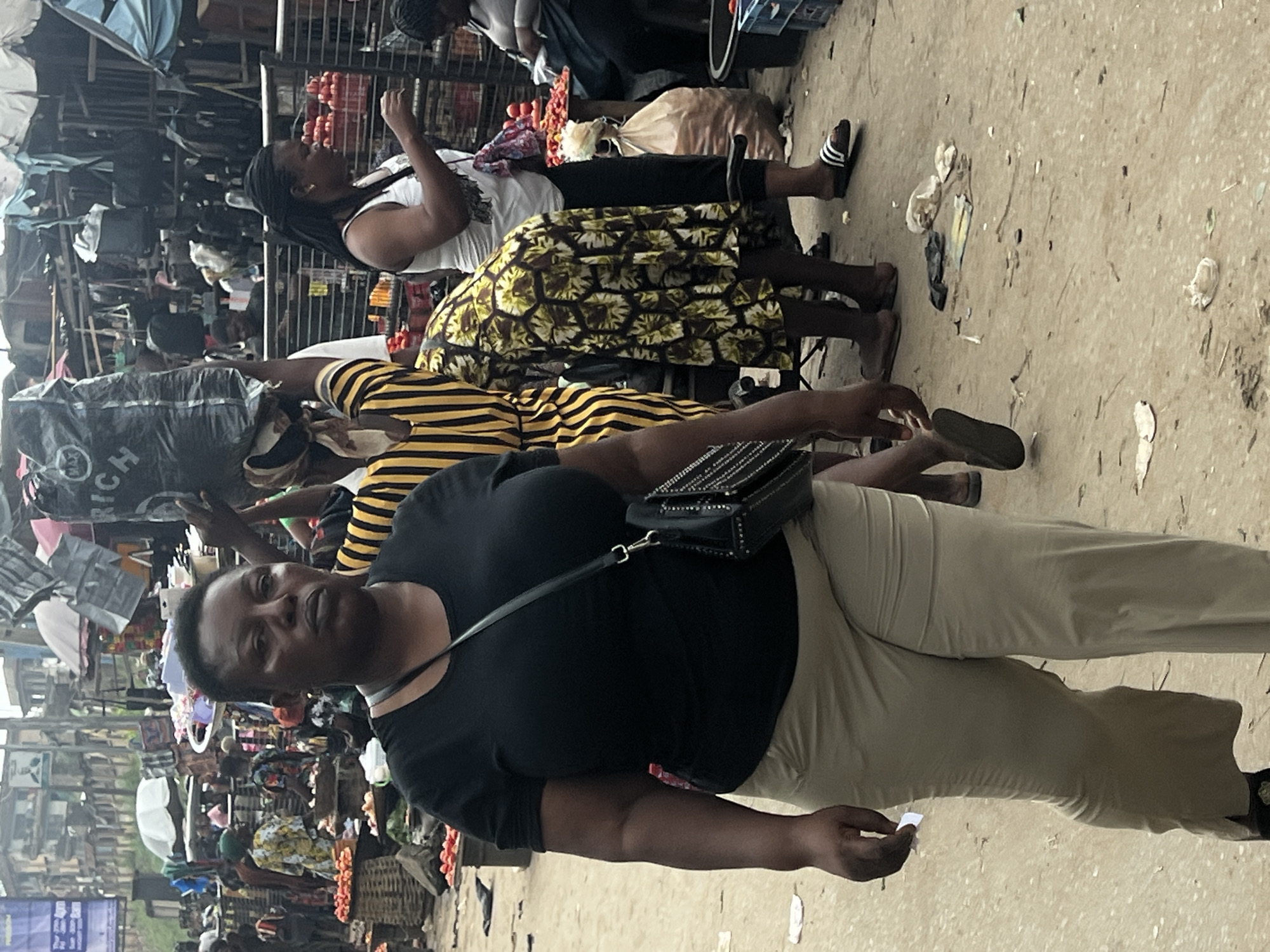 The image captures a street scene in Nkpor, Nigeria, likely a market environment. In the foreground, a Black woman walks towards the camera. She has dark skin, short dark hair, and is wearing a black t-shirt and tan pants. A black purse with metal studs is slung across her body. To her right and slightly behind her, another Black woman in a white tank top and dark pants walks away from the camera. She has braided black hair and is wearing a gold earring. Further to the right, a woman in a yellow and black striped dress is partially visible, carrying a large black plastic bag on her head. Behind these individuals, the scene opens up to a bustling market with stalls and numerous people. Various goods are displayed, including baskets of red produce, possibly tomatoes. Clothing racks and bags are visible in the background, suggesting a vibrant commercial area. The ground is dusty and appears unpaved, with scattered debris. The overall impression is of a busy, open-air market in an urban setting.