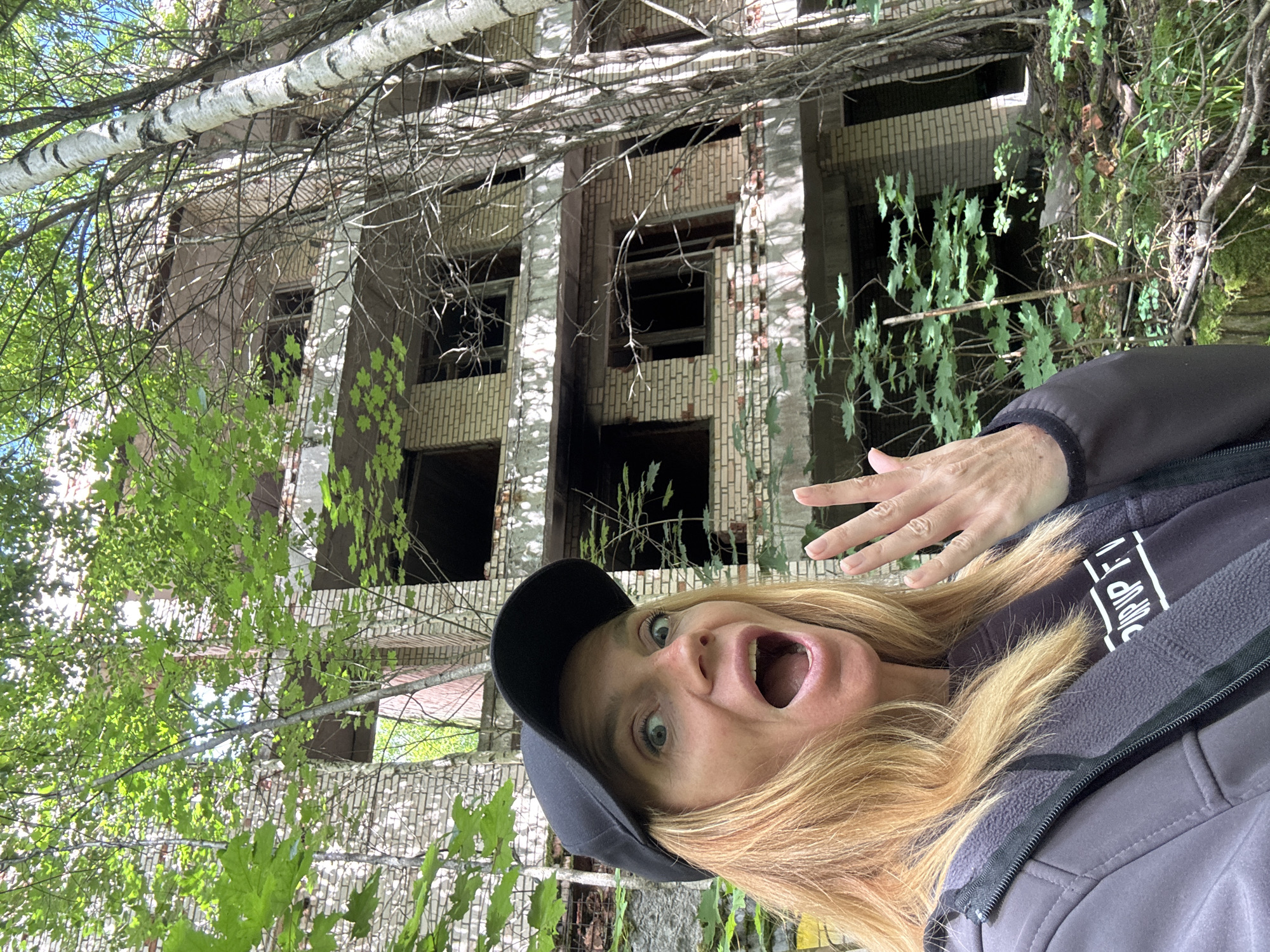 A woman wearing a black baseball cap and a dark jacket with the zipper pulled up to her neck is in the foreground. She has blonde hair and a surprised expression with her mouth open. Her right hand is raised, palm facing forward, with her fingers spread. She is wearing a dark t-shirt with white lettering that appears to say "POP UP FI". The background features a dilapidated multi-story building constructed of light-colored bricks, with numerous broken windows and decaying concrete floors. Trees with green leaves and a tall, slender birch tree with white bark are growing around and in front of the building, indicating an overgrown, abandoned environment. The ground is covered with debris, moss, and vegetation. The overall scene suggests an abandoned urban or industrial setting in Interkompleks, Russia.
