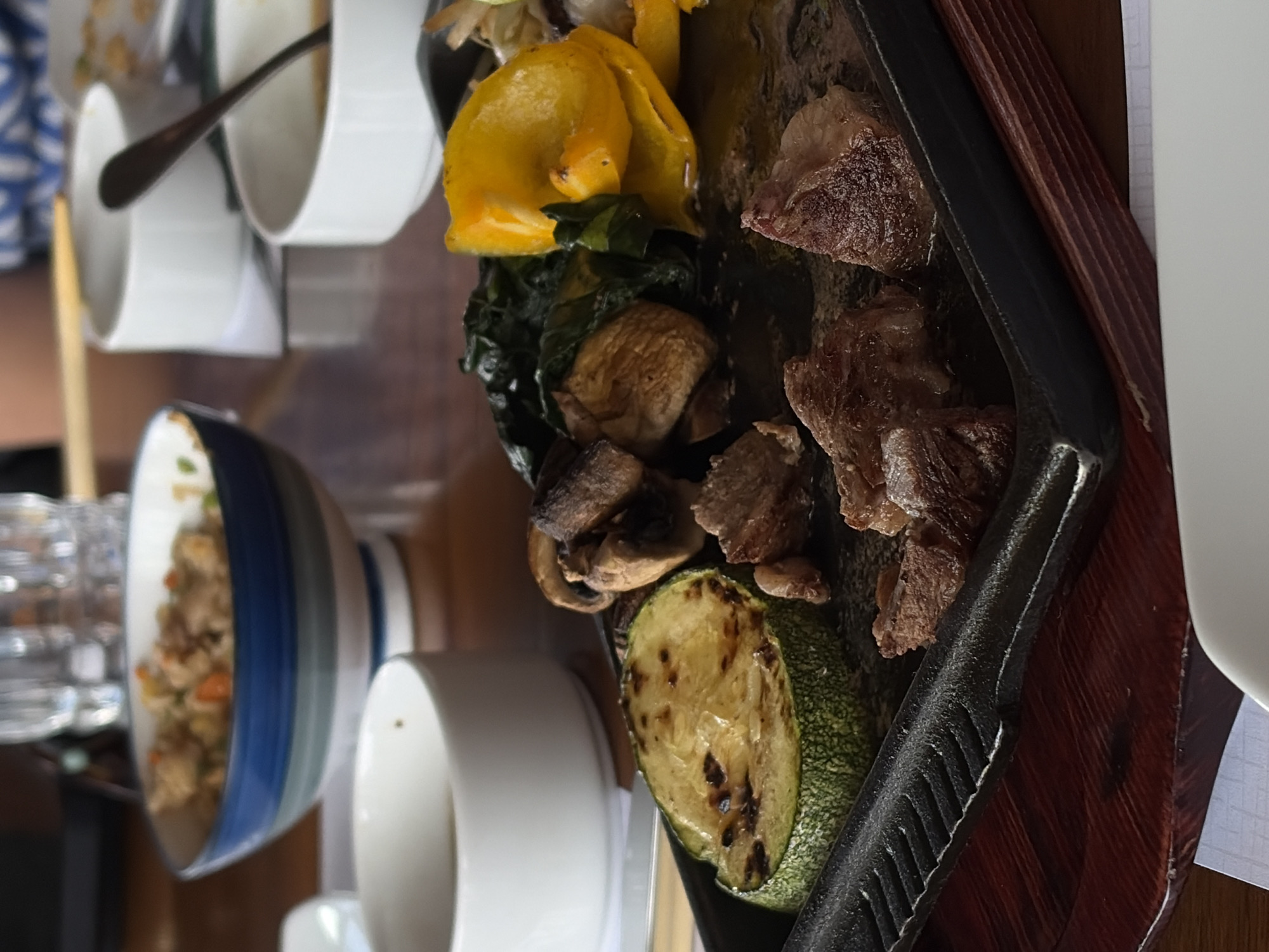 This image shows a meal setting at a restaurant, though the exact location in the city of Unknown, Unknown is unknown. Visible on the right side of the frame is a rectangular iron griddle with seared steak pieces, grilled zucchini, sautéed mushrooms, spinach, and yellow peppers. Next to the griddle are stacks of white teacups. To the left, there's a bowl with rice. In the background, a glass, chopsticks, and additional tableware can be discerned. The setting appears to be a casual dining environment.