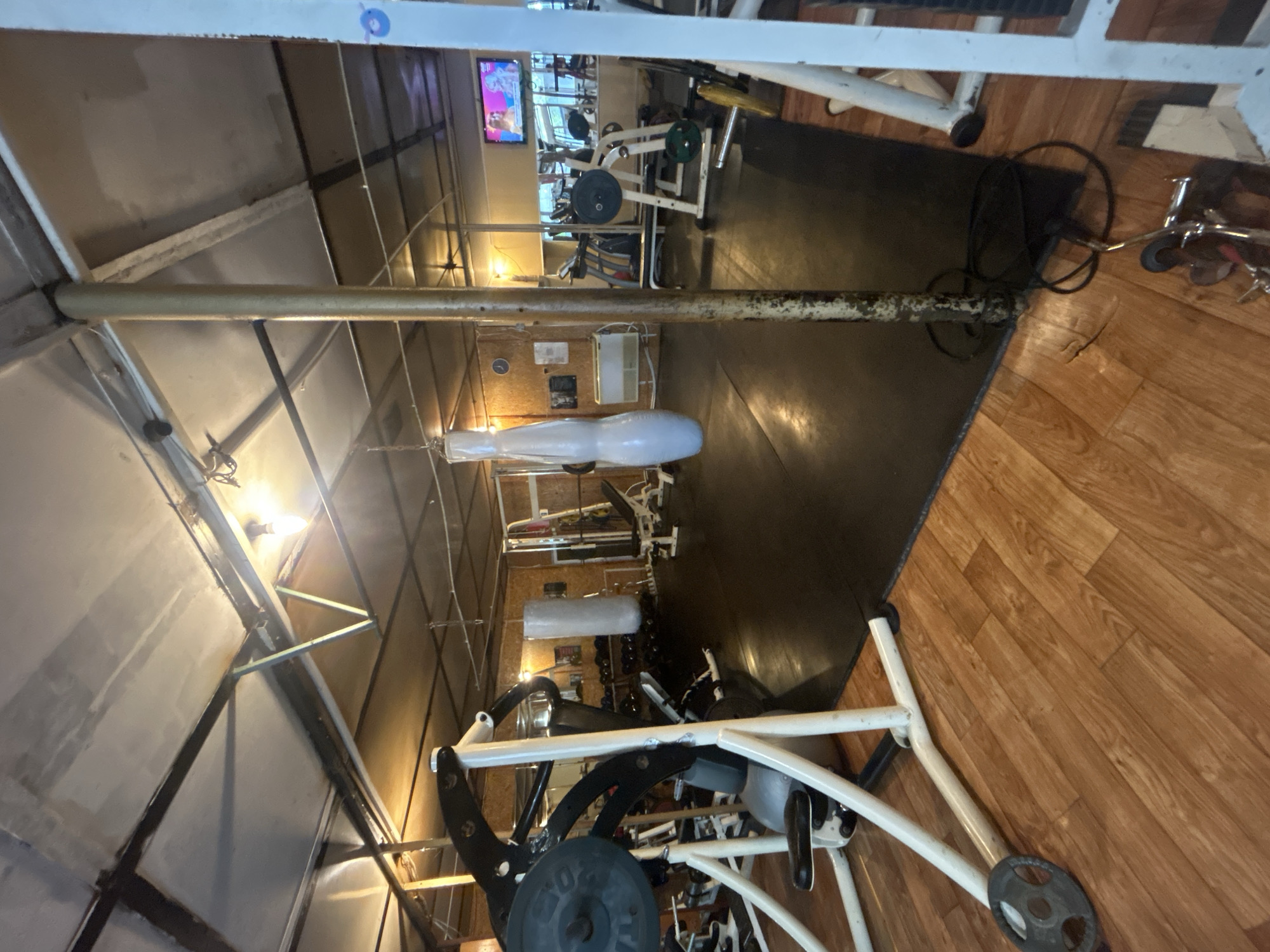 The image shows the interior of a gym. There are various weight machines and equipment scattered throughout the space, some of which are white-framed with black weights. A television screen is visible in the distance. The flooring transitions from dark matting in the center to wood-patterned flooring on one side. The ceiling appears to be constructed from corrugated metal sheets with metal supports. The overall lighting is somewhat dim. Elhovo, Bulgaria is not mentioned in the image.