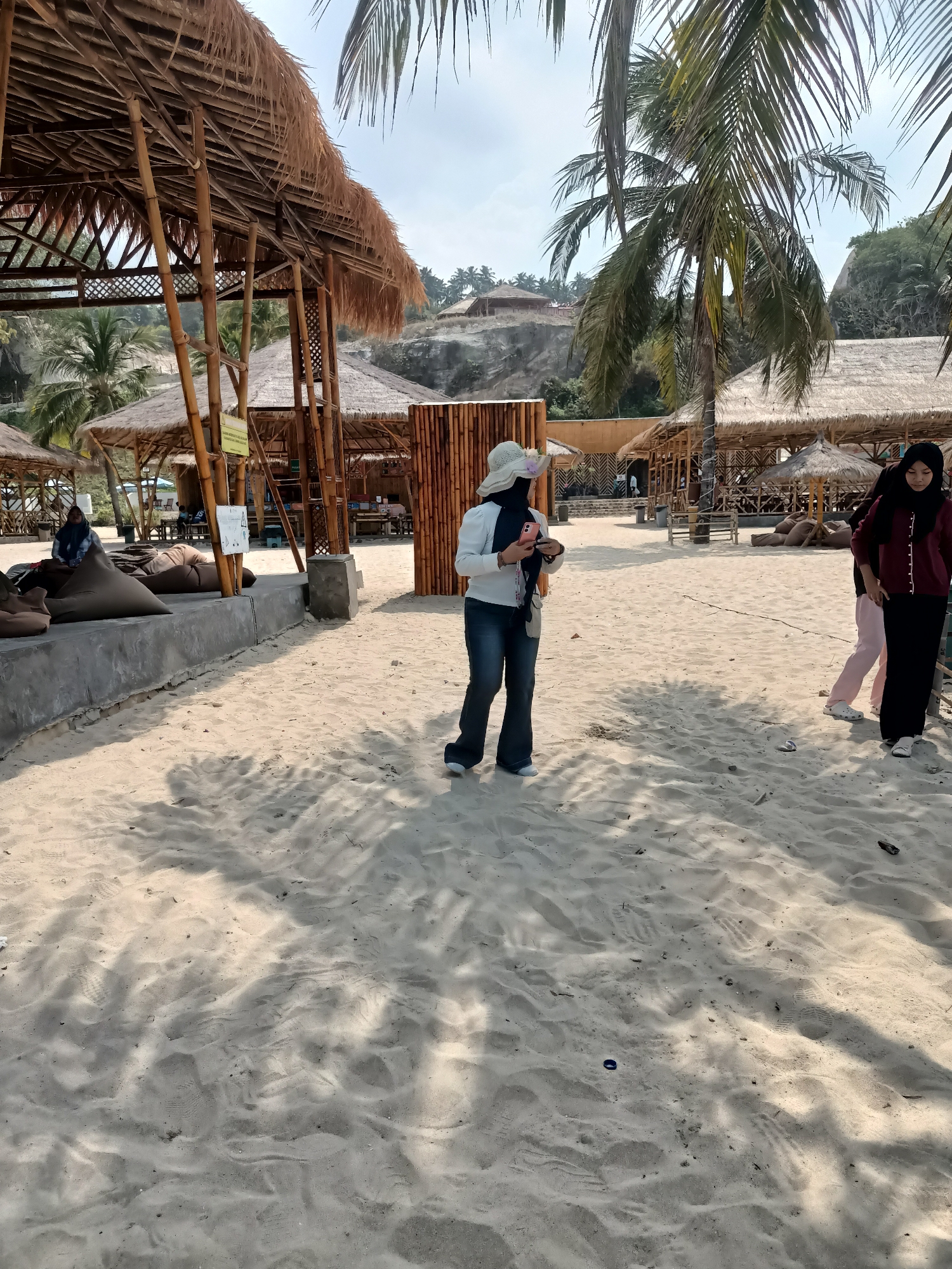 A sunny day unfolds at a beachside establishment in Bulok, Indonesia, featuring open-air bamboo structures with thatched roofs offering shade. In the foreground, white sand is imprinted with footprints and scattered with shadows from overhead foliage. A woman, wearing a white hat, a light-colored top, and jeans, stands facing left, holding a phone. To her right, another woman in a black hijab and maroon cardigan stands facing forward, while a third person in a pink outfit is partially visible behind her. Beanbag chairs are arranged on a concrete platform under one of the structures, where another individual is seated. The background shows more bamboo structures, palm trees, and a hillside with a few buildings.