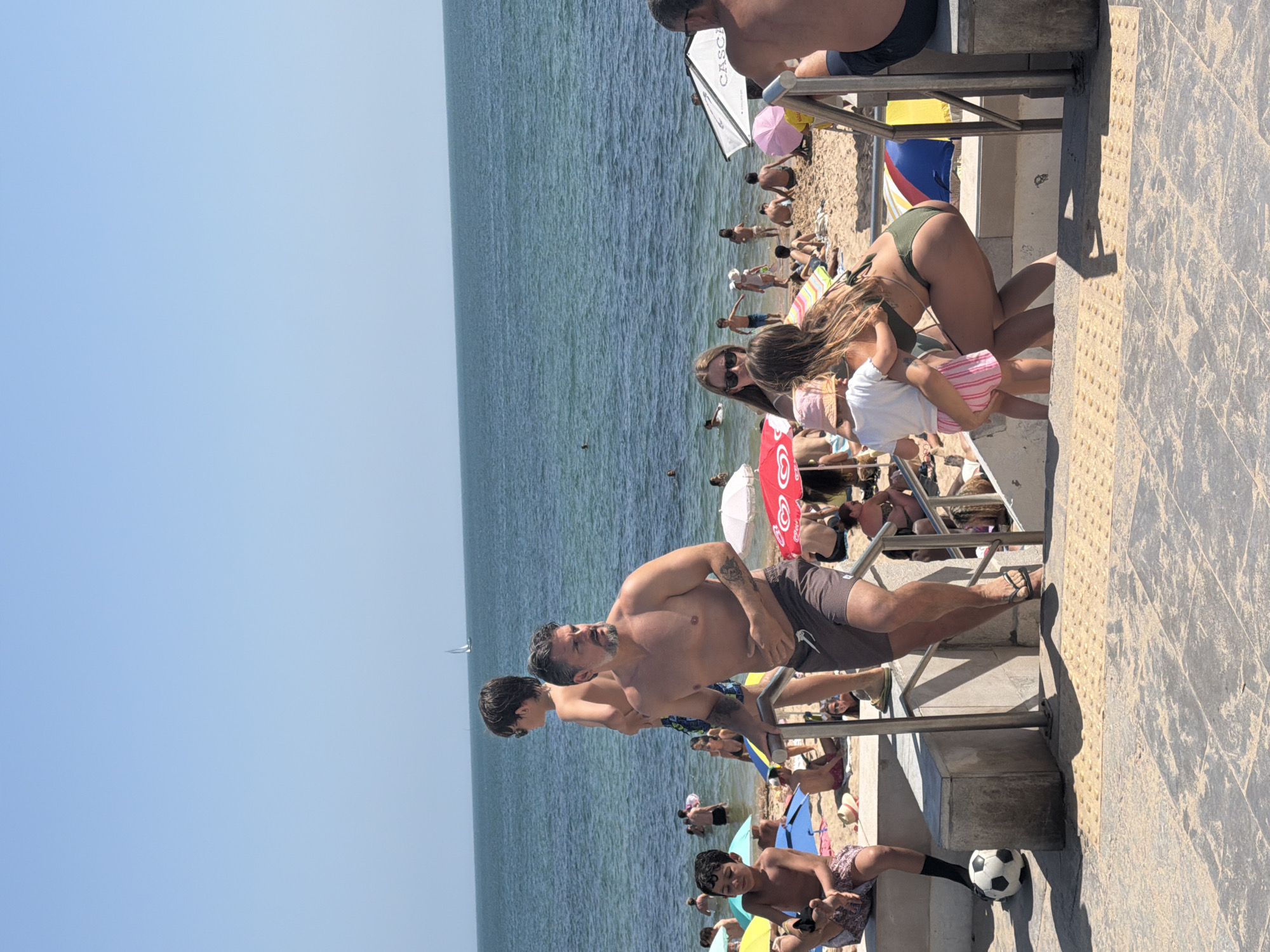 A busy scene unfolds on a sunny day at Praia da Rainha, Cascais, Portugal, with a crowd of people enjoying the beach and the ocean. In the foreground, a man with a beard and a young boy are descending stone stairs with metal railings. The man is wearing swimming shorts, and the boy is barefoot. A soccer ball rests near the boy.
On the beach, various individuals are spread out, some sunbathing, others wading in the water. Beach umbrellas in shades of red, white, pink, and blue dot the sandy shoreline. In the middle ground, a man in swim trunks is standing, looking out at the sea, while a young boy in patterned shorts sits on a wall. Further back, people are visible in the water, and a sailboat can be seen in the distance on the blue ocean under a clear sky. The overall impression is of a typical summer day at a popular beach location.