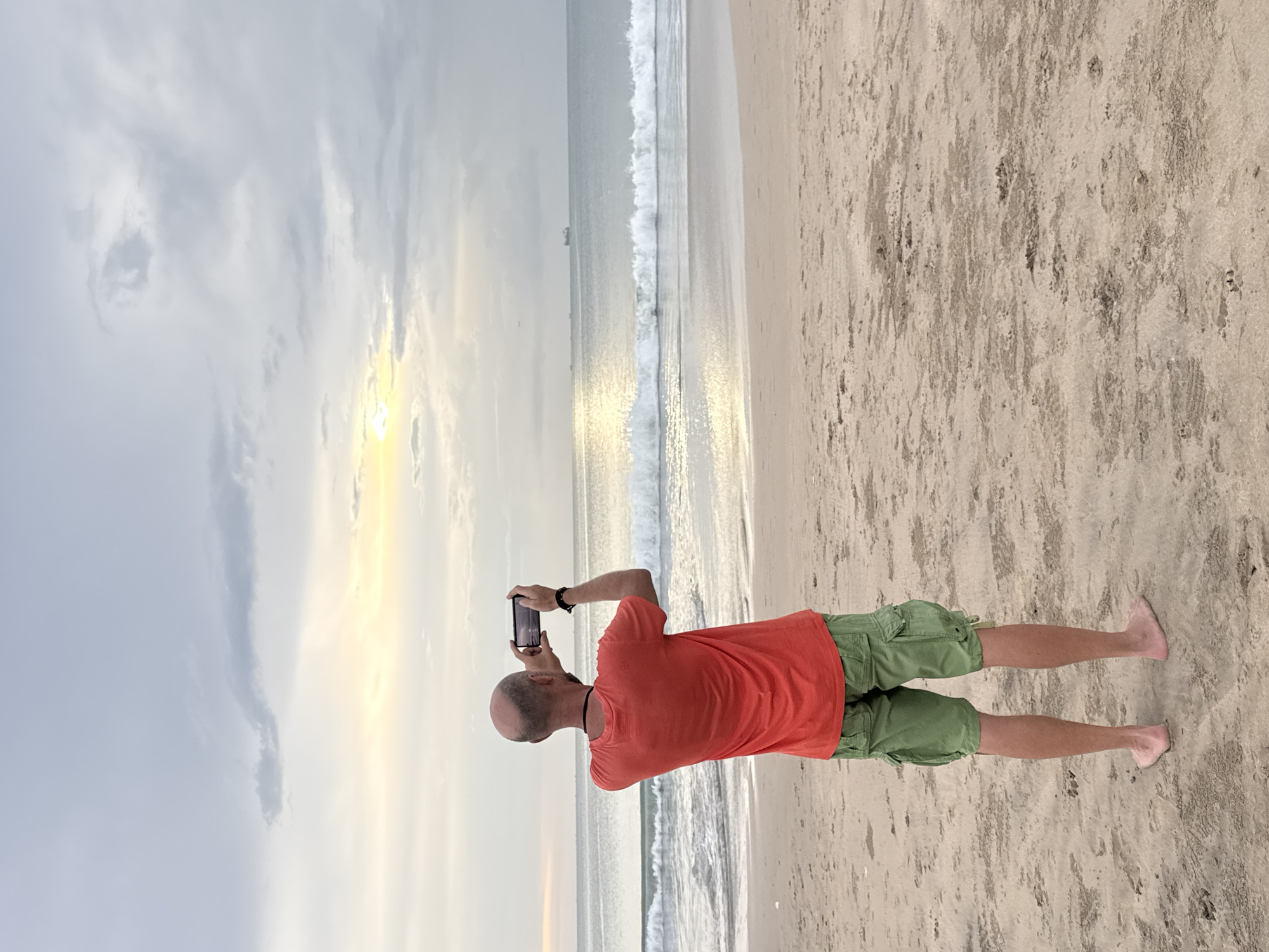 A photograph taken on a beach in Kuta Selatan, Indonesia, shows a bald man standing on the sand facing the ocean and taking a photo with his mobile phone. The man is wearing an orange t-shirt and light green cargo shorts. The ocean waves are visible, with a line of white foam. The sky is partly cloudy, with the sun reflecting off the water. The sand is light-colored.