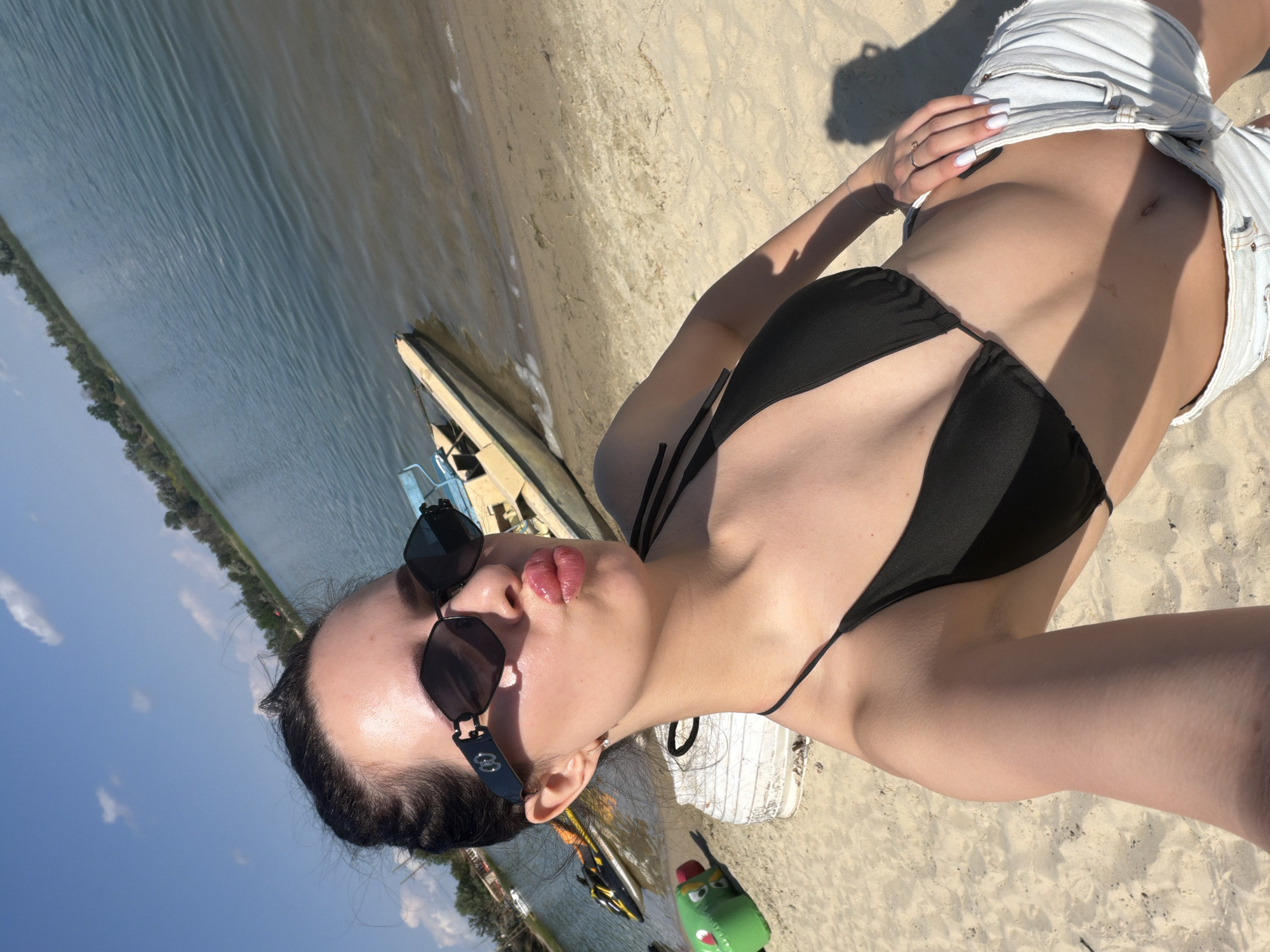 A young woman with dark hair pulled back in a bun, wearing black sunglasses and a black bikini top with denim shorts, poses for a selfie on a sandy beach next to a body of water. She is puckering her lips, and her left hand is resting on her hip. In the background, a shallow boat is partially submerged in the water near the shore, and a jet ski is visible further back. The environment is sunny with a clear blue sky and some scattered clouds. The overall scene suggests a recreational setting at a lake or similar body of water.