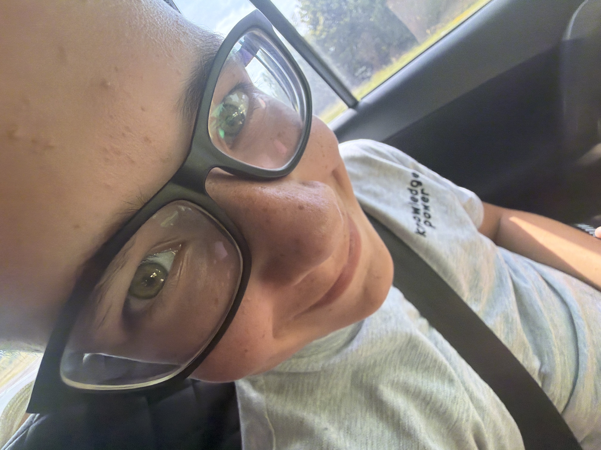 A low-angle, close-up selfie captures a person wearing black-rimmed glasses and a grey t-shirt with the text "knowledge power" on it, seemingly inside a car. The person's green eye and part of their nose and mouth are visible through the left lens of the glasses, reflecting the outdoor environment. Their head is tilted, and they are smiling slightly. The background is blurred, showing foliage and the sky, suggesting an outdoor setting. The seatbelt is clearly visible across the person's chest. The image quality is grainy, and the lighting suggests it was taken during the daytime. There is no visible indication of the location being Kruglovka, Russia.