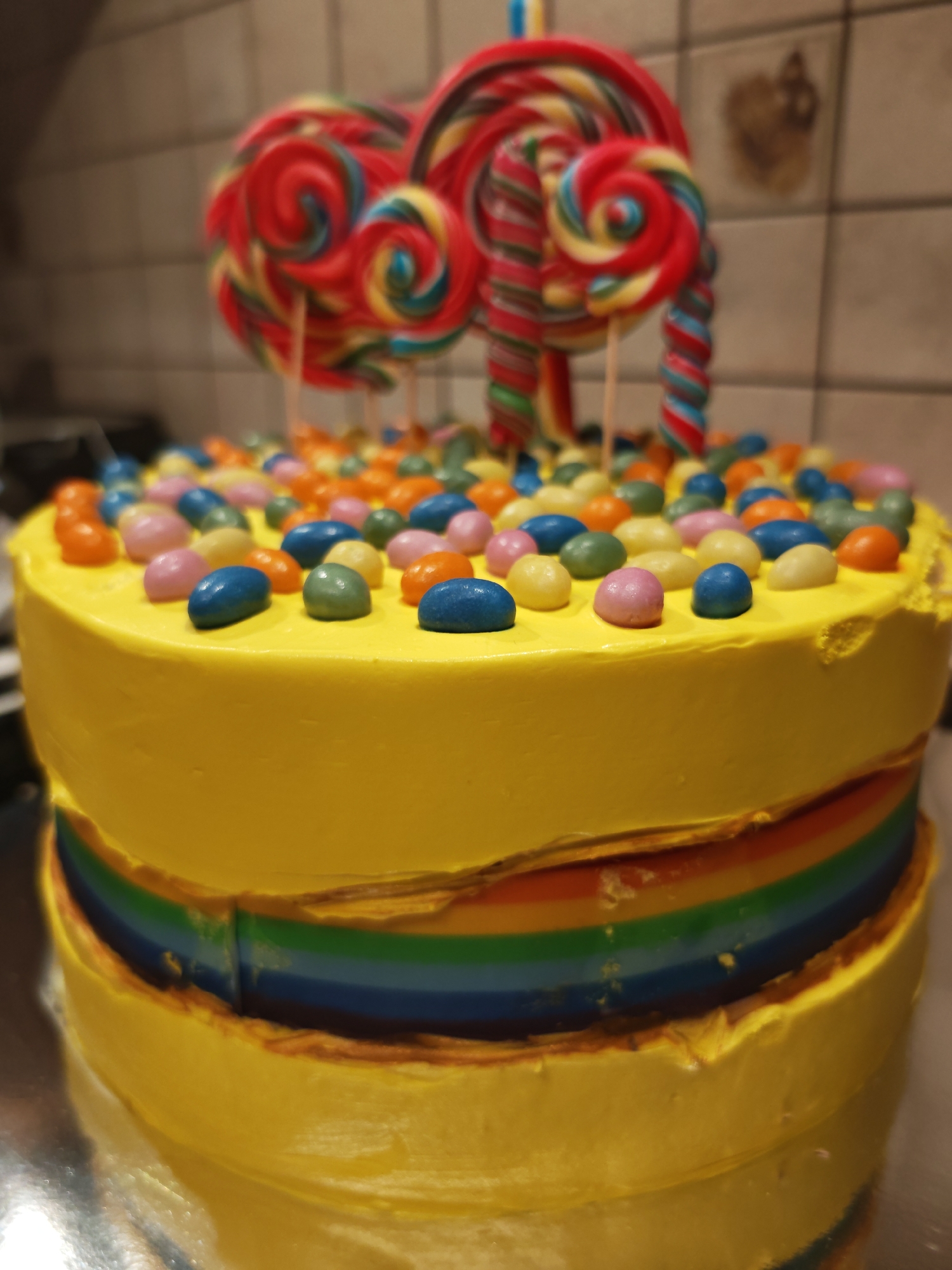 A colorful cake, decorated with colorful candies and lollipops, sits on a reflective surface. The cake has a yellow frosting and features rainbow layers visible around its perimeter. The background shows a tiled wall. The image does not contain any information that can be used to determine its location in Sofia, Bulgaria.