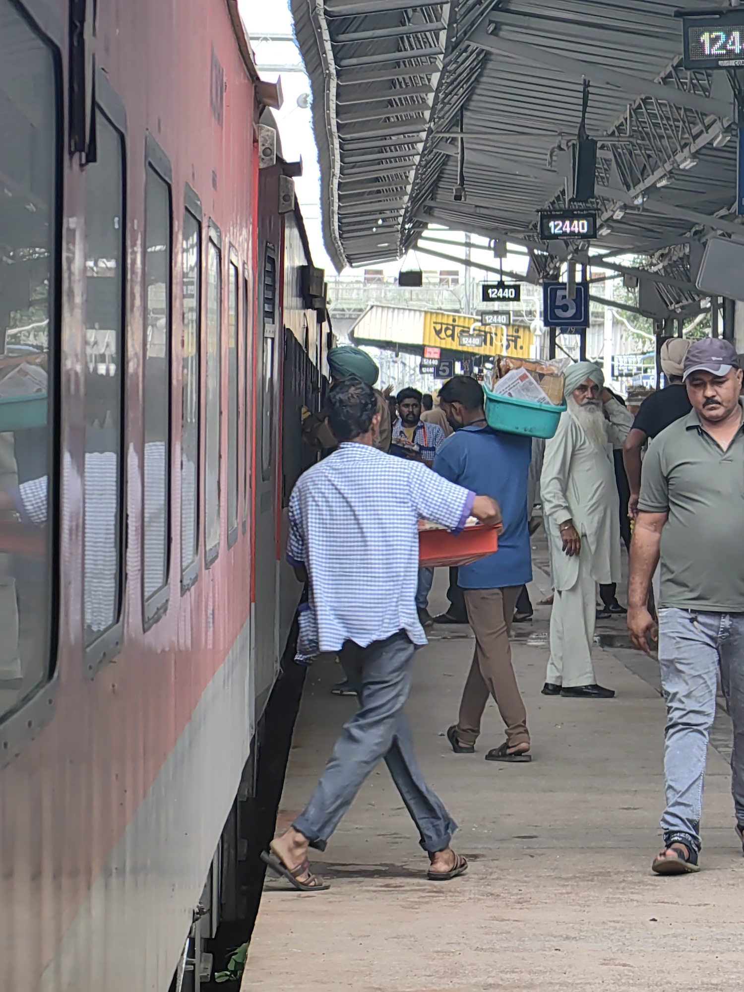 The image depicts a busy train platform at Khandwa Junction railway station in Khandwa, India. A red passenger train is parked alongside the platform. In the foreground, a man in a blue and white checkered shirt is walking away from the train, carrying a red tray with some items. To his right, another man in a blue shirt is carrying a green basket on his shoulder, filled with various goods, including newspapers and what appear to be packaged food items. Further in the background, an elderly man with a long white beard and a turban is standing and looking towards the train. Other people are visible on the platform, some waiting, some walking. Overhead, the station's metal roof structure is prominent, with digital display boards showing train information. The platform is made of concrete. The overall scene suggests normal activity at a train station during the day.