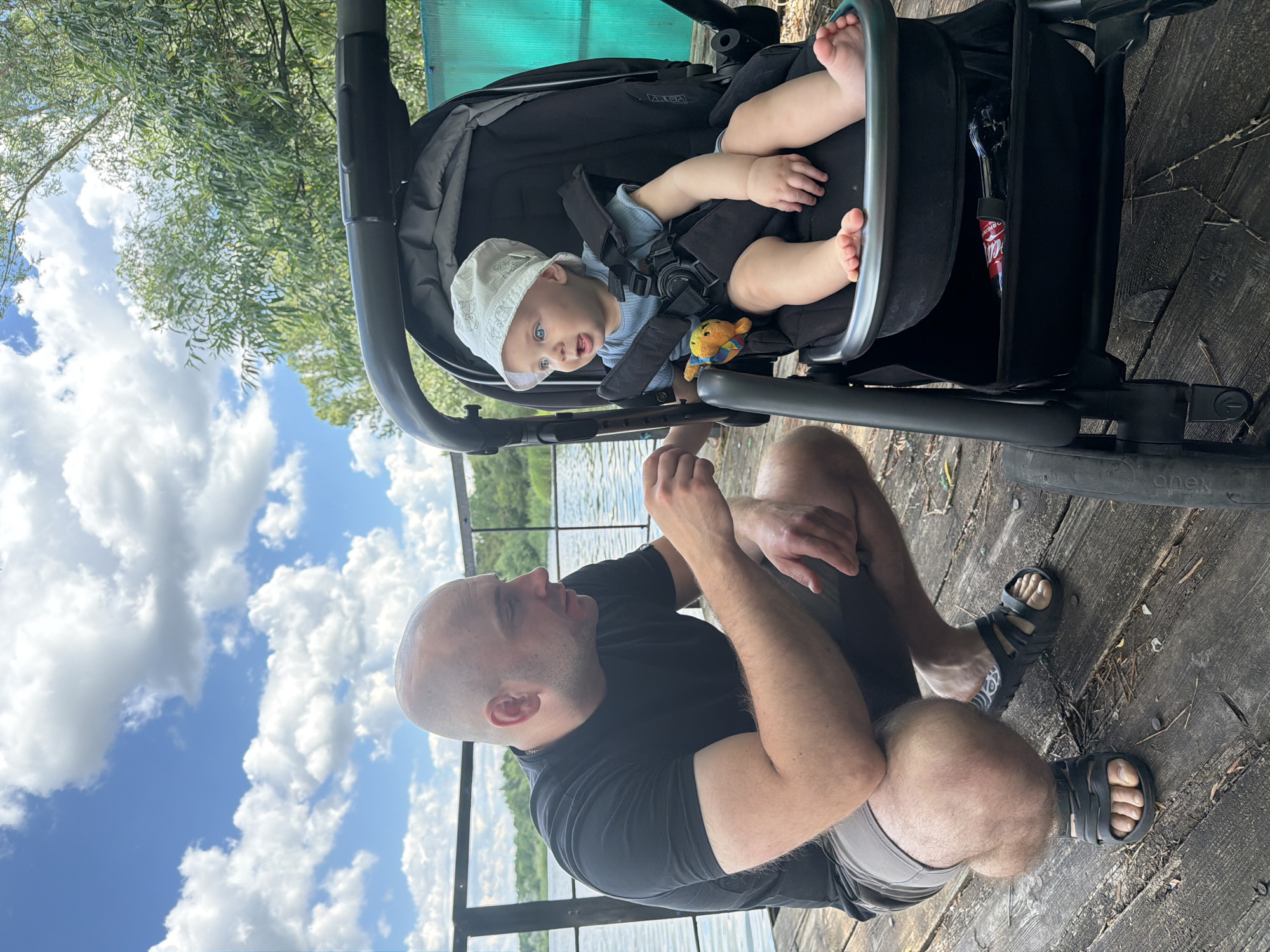 A bald man in a black t-shirt and gray shorts squats on a wooden dock next to a baby stroller. The stroller contains a baby wearing a white hat and a blue shirt. The baby is looking to its right. In the background, there is a body of water and trees. The sky is blue with white clouds. The scene appears to be in Lozuvatka, Ukraine.