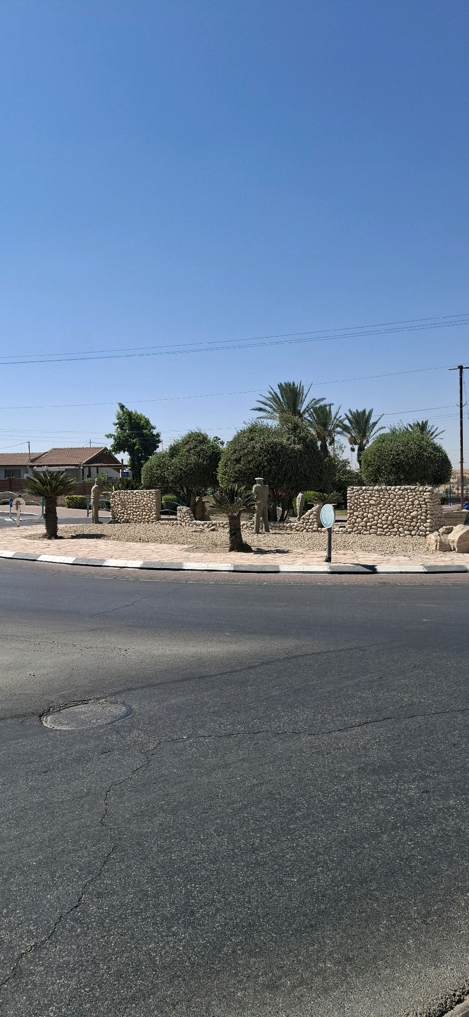 The image shows a street scene in Dimona, Israel. In the foreground, there is a cracked asphalt road. In the background, there is a circular garden area with low stone walls, sculpted bushes, palm trees, and statues of people. One statue appears to be a soldier. Behind the garden, there are buildings, including a house with a brown roof, and palm trees. The sky is clear and blue. The scene is illuminated by bright sunlight, suggesting it is daytime.