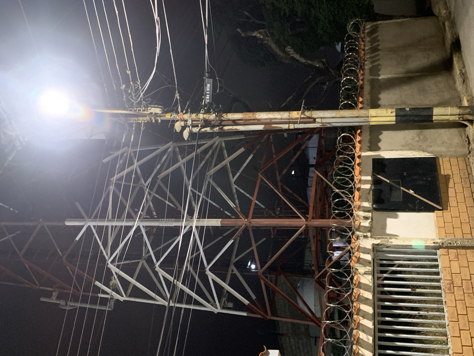 The image displays a nighttime street scene in Santa Ana, Venezuela, dominated by a large metallic lattice tower and a brightly illuminated utility pole. The central lattice tower is a multi-story structure, exhibiting sections painted white and rust-red/brown. Visible within its lower framework are a cylindrical tank-like object and a small light source. Multiple overhead utility lines cross horizontally through the tower's mid-section.
To the right of the tower, a weathered utility pole supports numerous electrical and communication cables. A potent white street light fixture at the top of this pole emits significant light, creating a prominent lens flare. A black rectangular sign marked "CSI T 300" is affixed to the wiring bundle below the light. The lower section of the utility pole is painted with distinct black and yellow stripes.
In the foreground, a security fence separates the viewer from the property where the tower is located. This fence is constructed with a base of light brown bricks, topped by a concrete wall featuring terracotta-style coping. Coiled razor wire is installed along the entire top perimeter of the concrete wall. A section of a metal bar fence is visible to the left. Behind the fence, a dark building with a black door and dense vegetation are discernible against the dark background. No individuals are present in the scene.