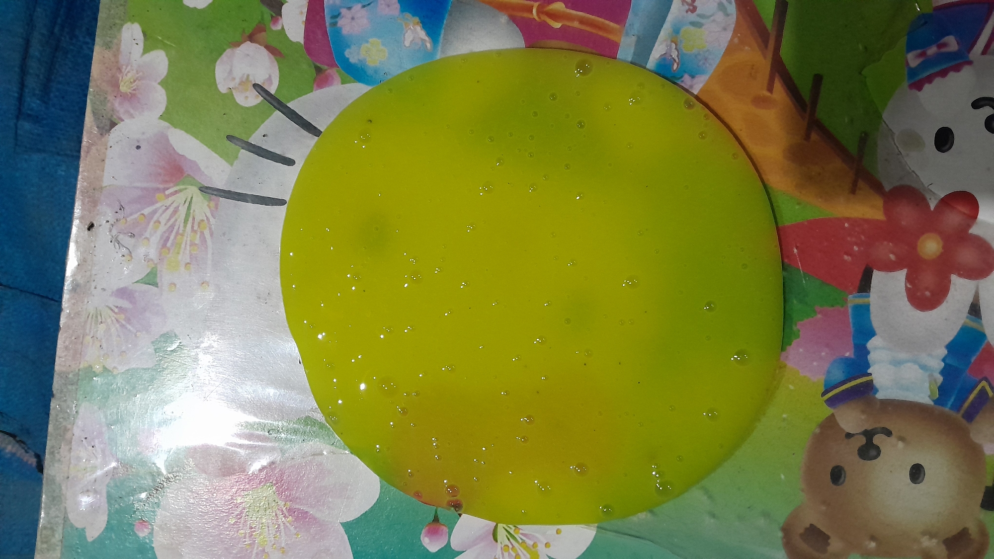 A close-up overhead shot shows a circular blob of bright green slime with small air bubbles on a surface with a colorful print. The print depicts light pink and white cherry blossoms against a teal background in the upper portion of the image. In the lower portion, the print transitions to feature cartoon characters, including a brown teddy bear in a blue and gold uniform, a red flower, and the face of a white rabbit. The slime is glossy and reflective, with tiny white highlights scattered across its surface. The overall impression is of a playful or craft-related activity. The image is sourced from Sumberarum, Indonesia.