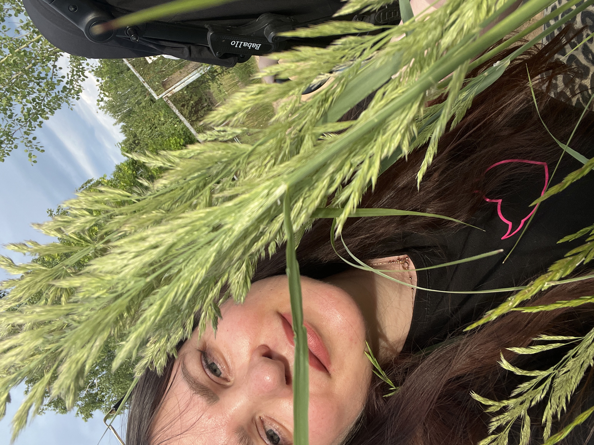 The image shows a woman with long, dark hair and fair skin in a close-up shot. She is partially obscured by tall green grass. She appears to be outdoors, with trees and a cloudy sky visible in the background. A black stroller is also visible to the right of the frame, with the brand "Baballo" printed on it. A metal fence is seen behind the grass and trees. The woman is wearing a black t-shirt with a neon pink heart emblem. She is also wearing a delicate necklace. The location is identified as Noviy, Russia.