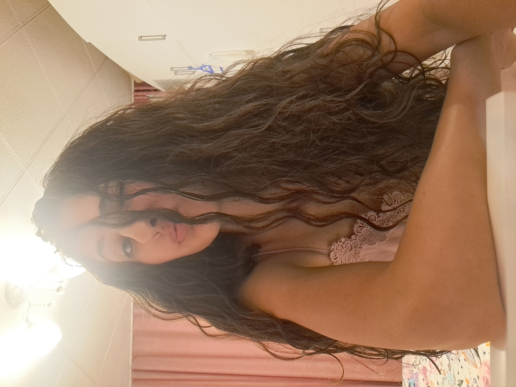 A woman with long, dark, curly hair leans forward, her right arm resting on a white surface. She is wearing a light pink lace camisole. Her face is partially obscured by her hair, but her lips are pursed. The background shows a pink curtain and a white closet. The image is taken indoors, likely in a bedroom. There is no indication of the city of Negovan, Bulgaria.